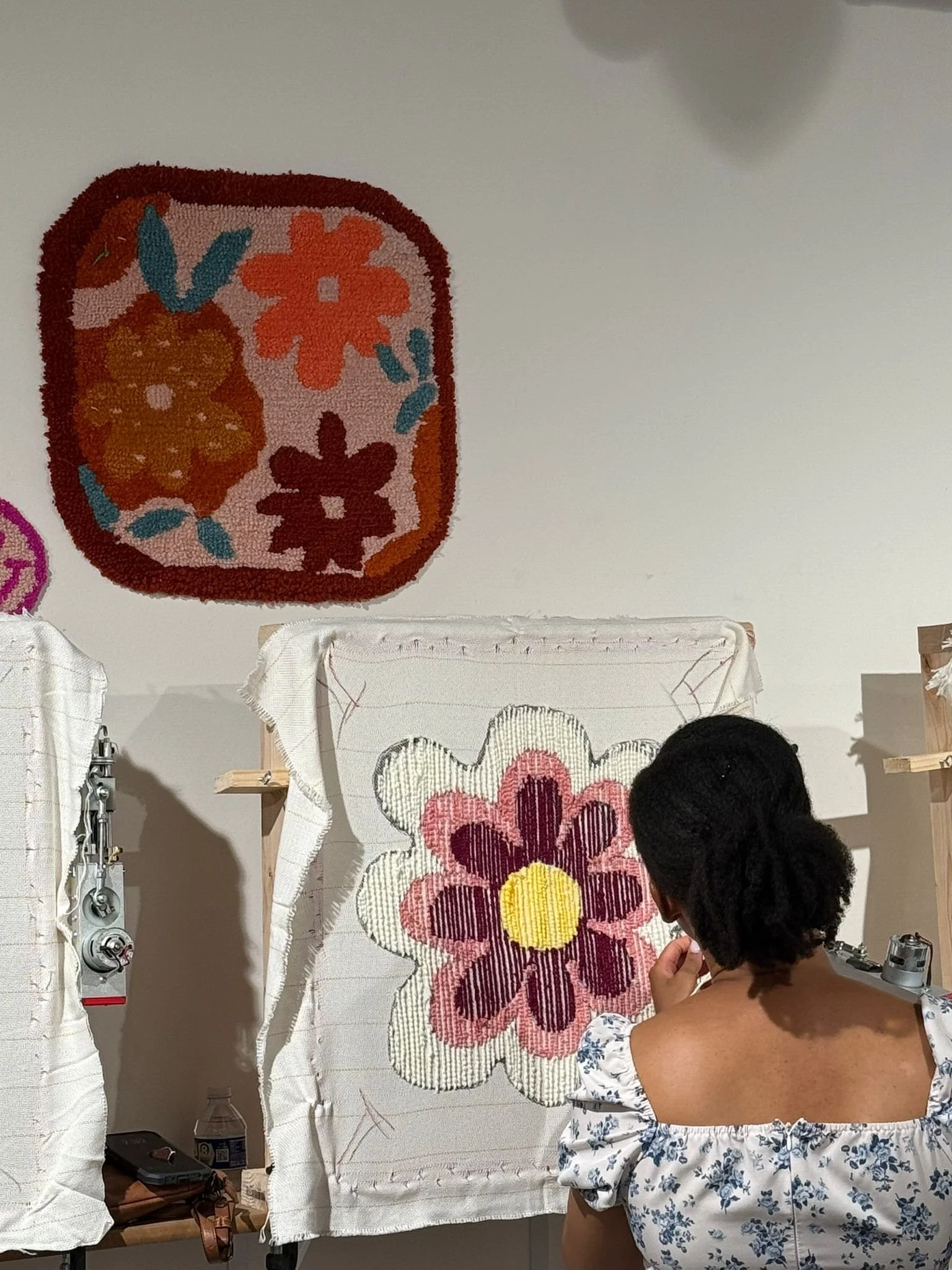 Woman working on floral needlepoint tapestry in an art studio, with a completed floral piece hanging above her.