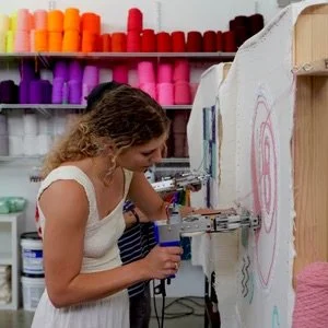Young woman using a drill to create art on a large white canvas in an art supply store with colorful yarn on shelves in the background.