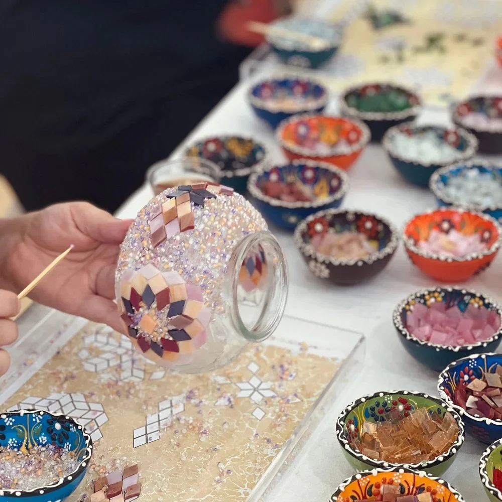 Person holding a mosaic vase with colorful tiles and beads with small bowls filled with mosaic pieces on a table.