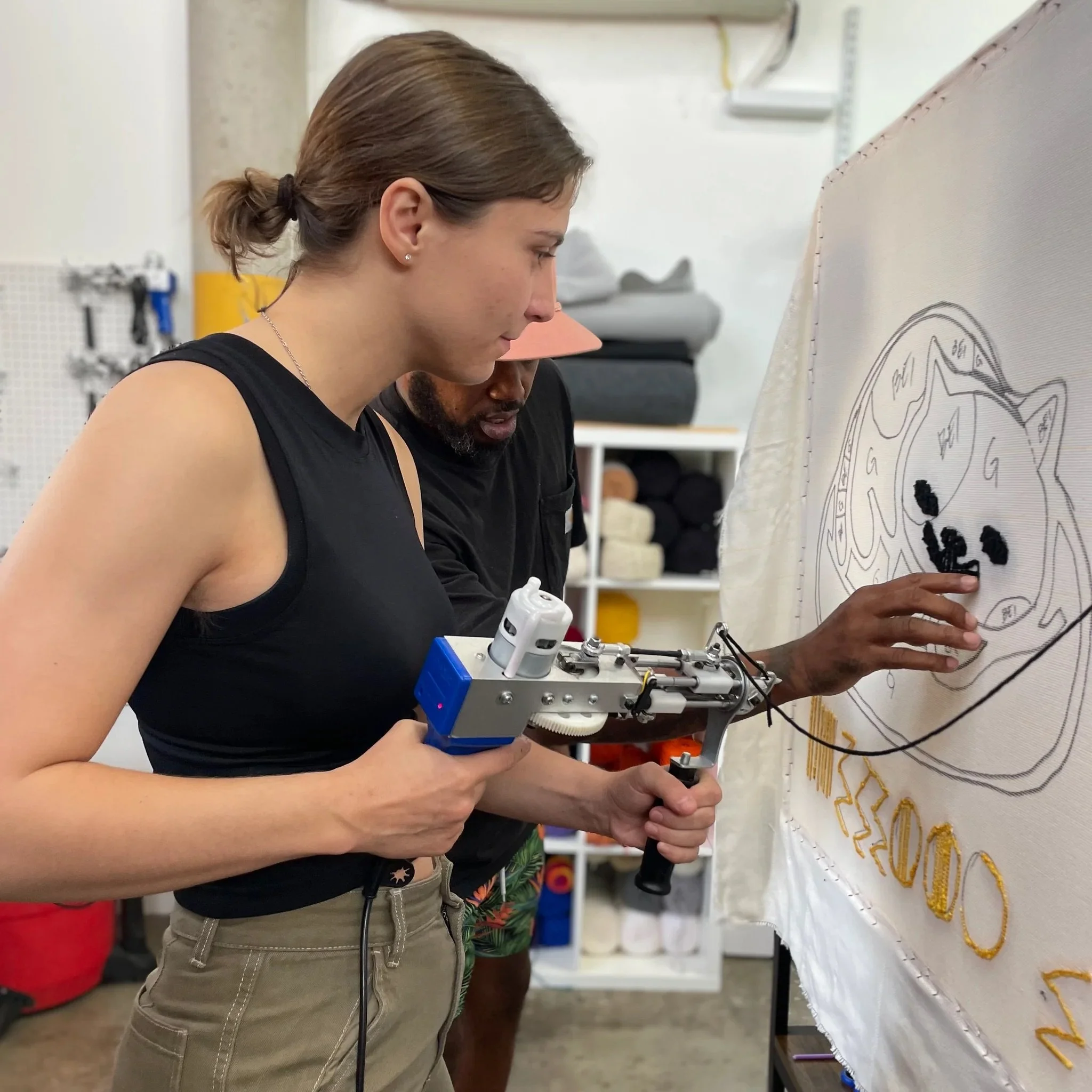 A woman operating a robotic device to embroider or add detail to a large fabric artwork of a lion's face, with a man observing nearby in a workshop setting.