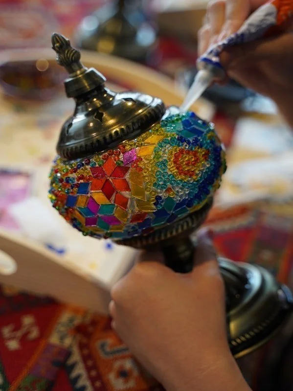 Person decorating a mosaic lamp with colorful glass pieces and beads, using a tool to apply adhesive.