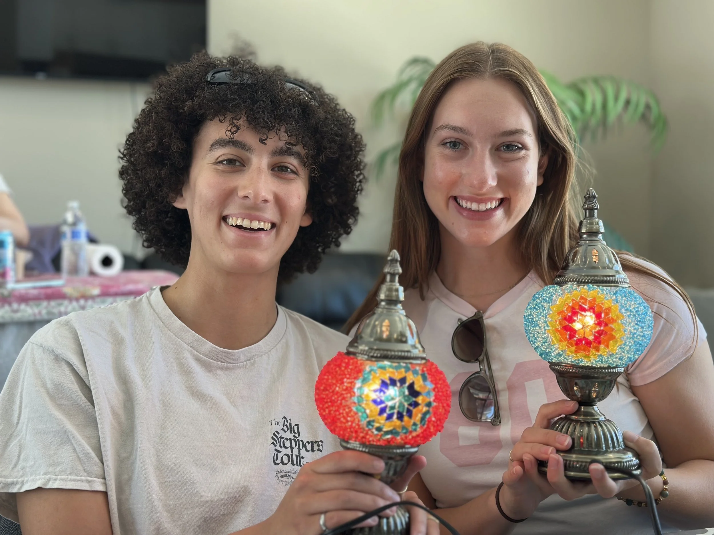 Two young women smiling and holding decorative mosaic lamps indoors.