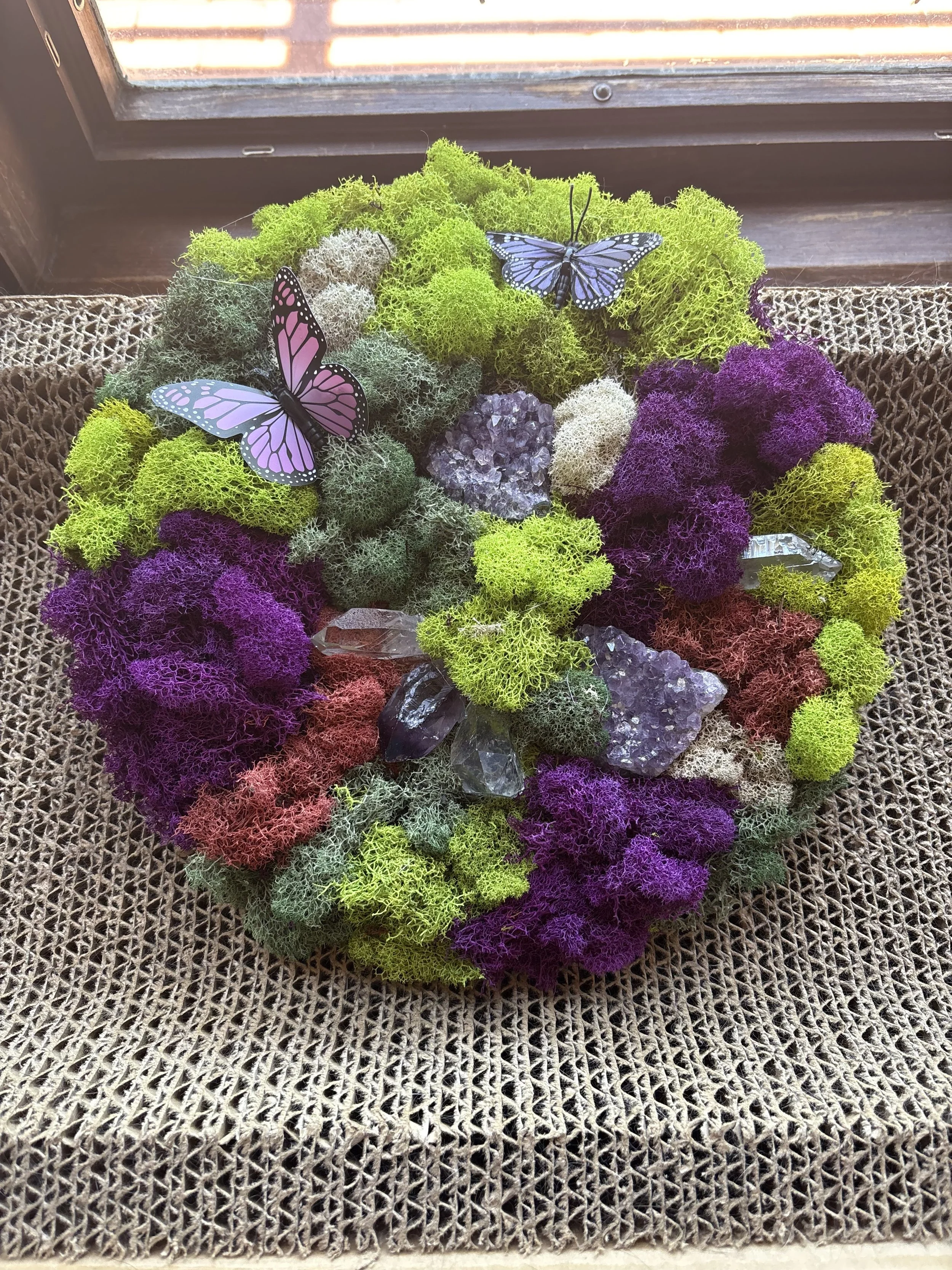 Colorful moss and crystals arranged in a circular pattern with butterfly decorations on a woven surface near a window.