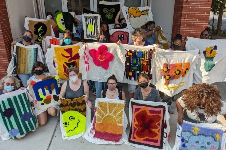 Group of people holding colorful, handmade woven and textile art pieces outside a building.