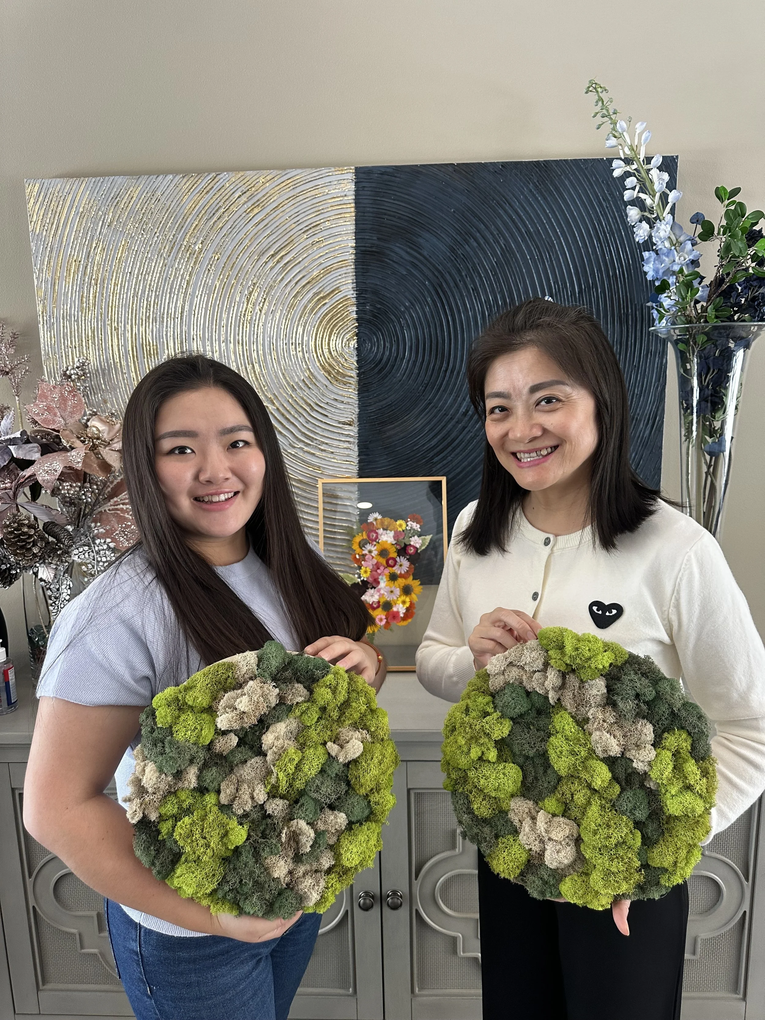 Two women standing indoors, each holding a large floral arrangement made of moss and succulents, smiling at the camera. Behind them is a decorative wall art and flowers.