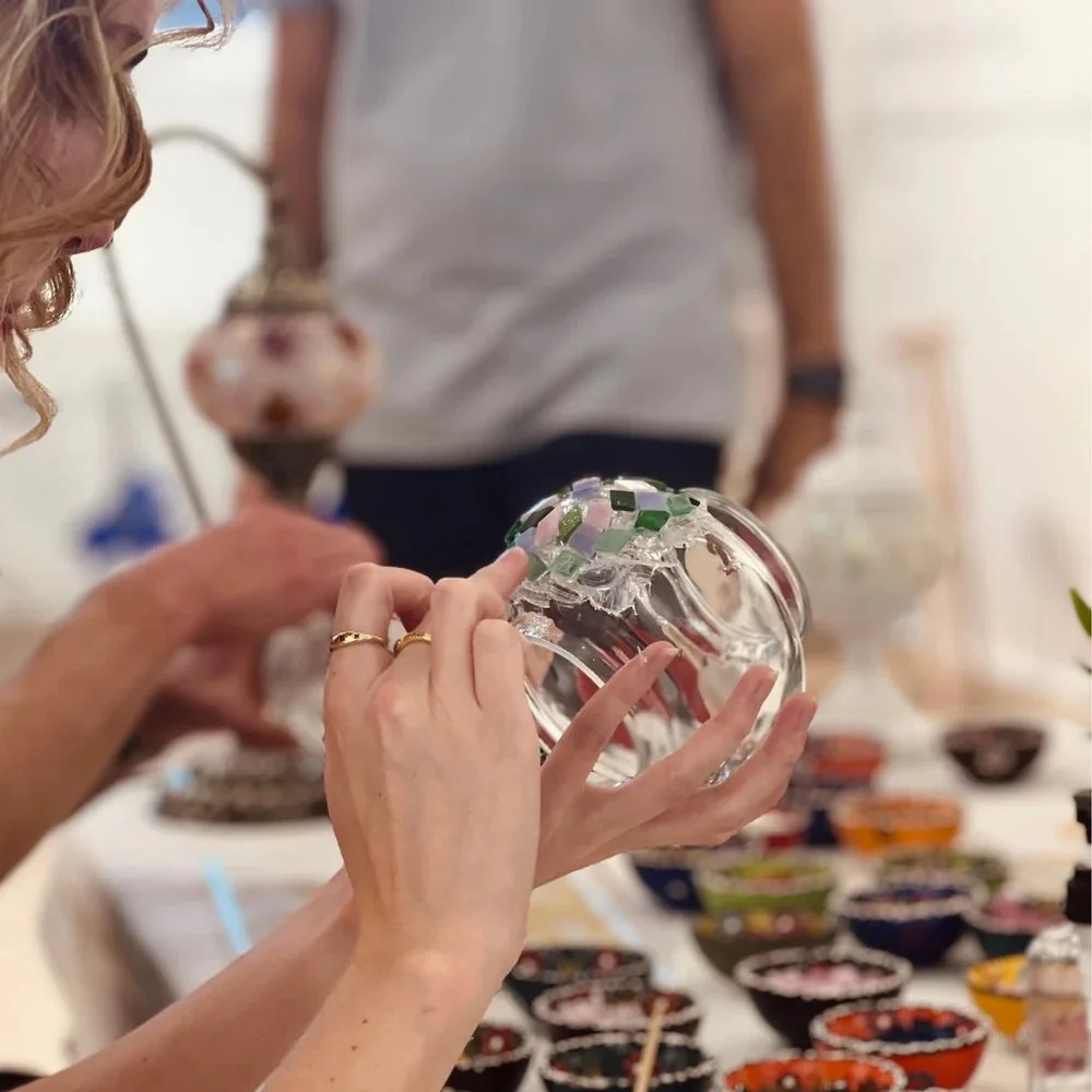 Person decorating a glass object with mosaic tiles, surrounded by small colorful bowls, in a crafting setting.