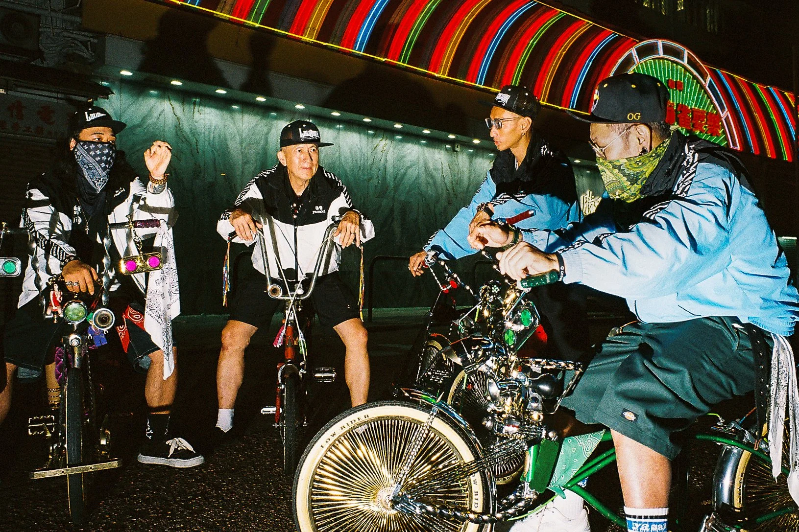 Four riders on custom lowrider bikes under neon signs on a Hong Kong street at night