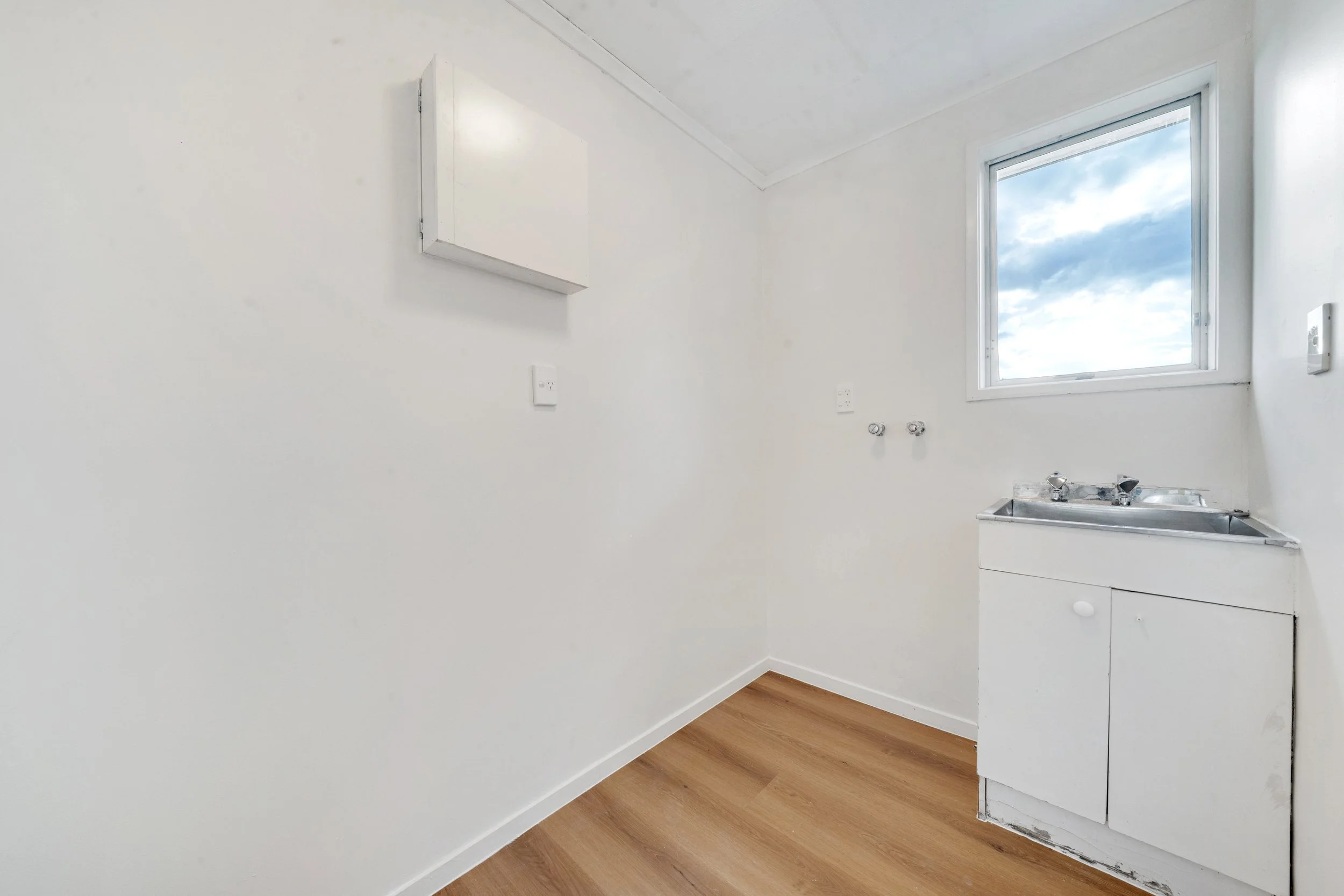Empty laundry room with white walls, a small window showing a cloudy sky, a white cabinet, and a metal sink with a wooden floor.