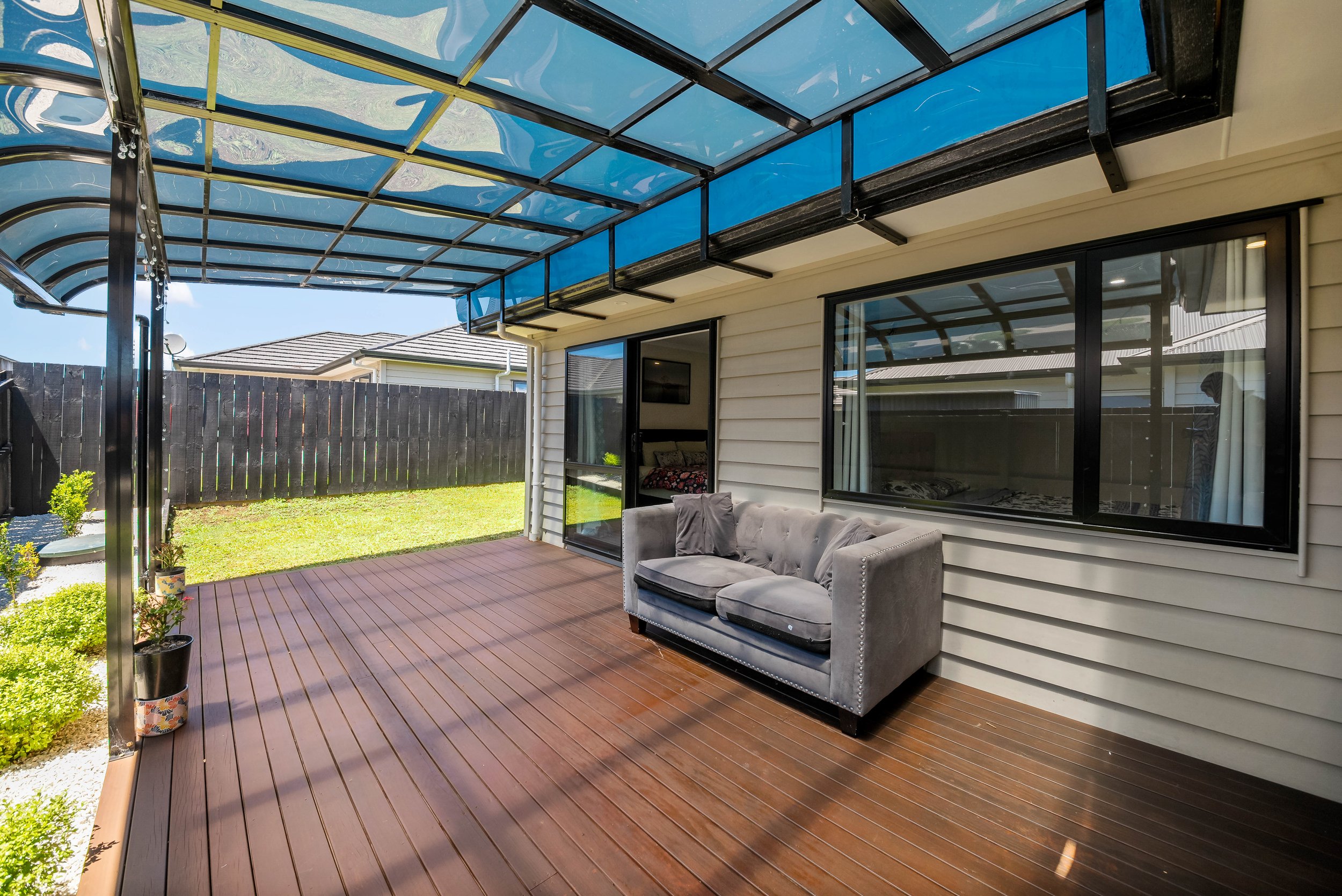 Backyard patio with wooden floor, gray outdoor sofa, potted plants, and a blue shade cover. Windows and sliding door lead into the house, fence in the background.