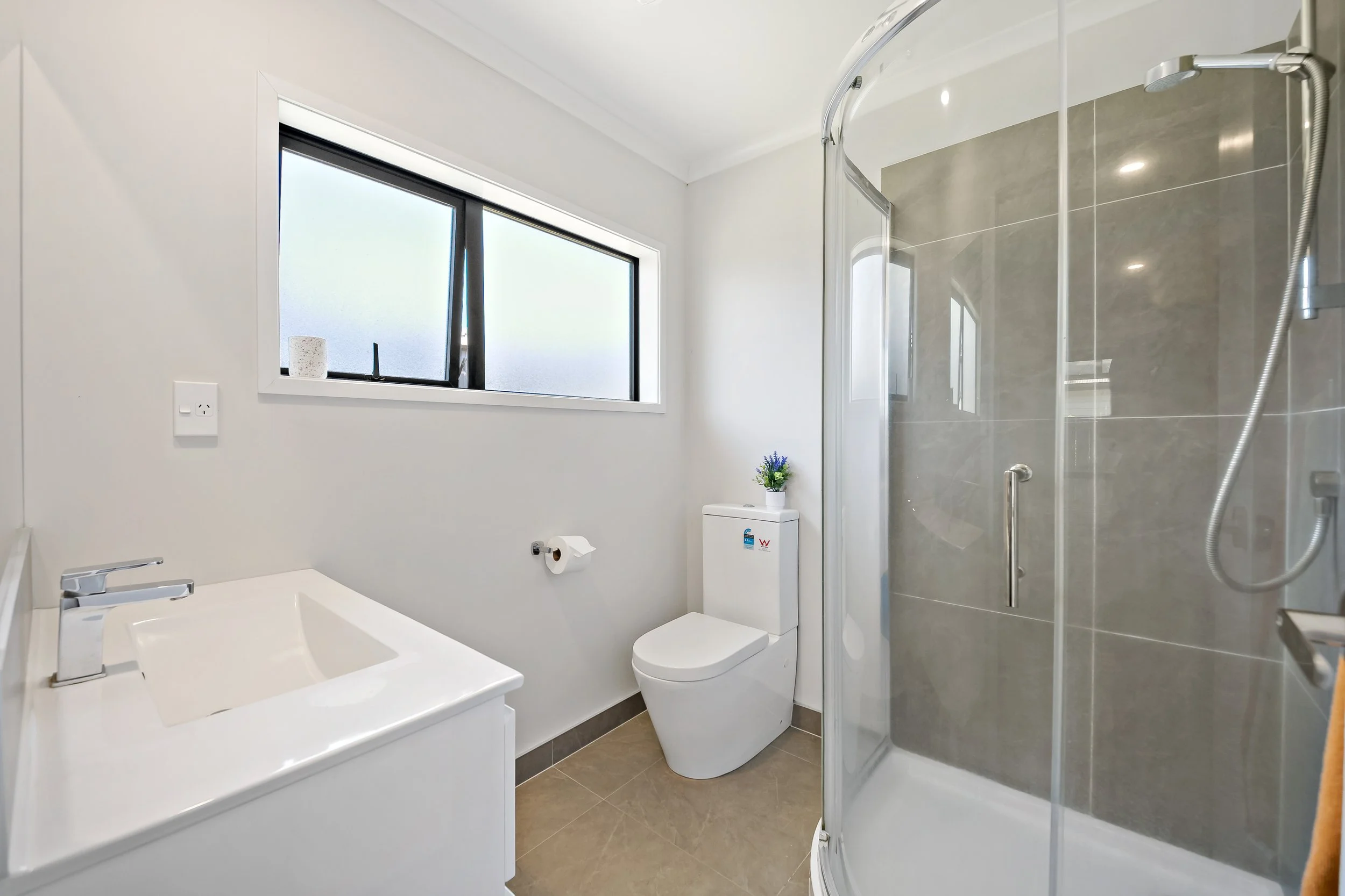 Bathroom with white walls, a window, a white toilet with a small plant on top, a corner shower with glass door, and a white vanity with a sink.