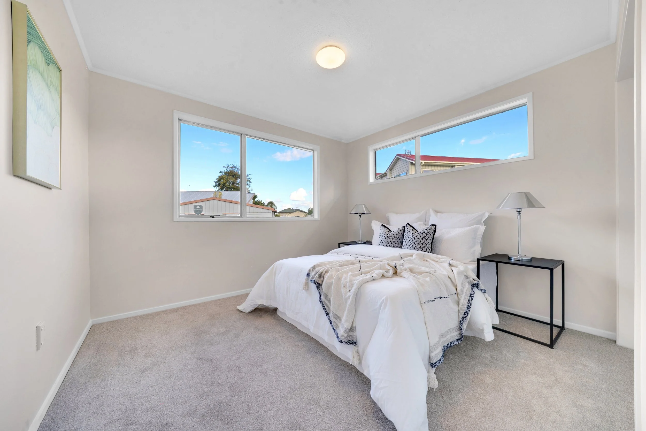 A bedroom with a bed, white bedding, black and white pillows, two bedside tables with lamps, beige carpet, and two windows showing a clear blue sky and neighboring houses.
