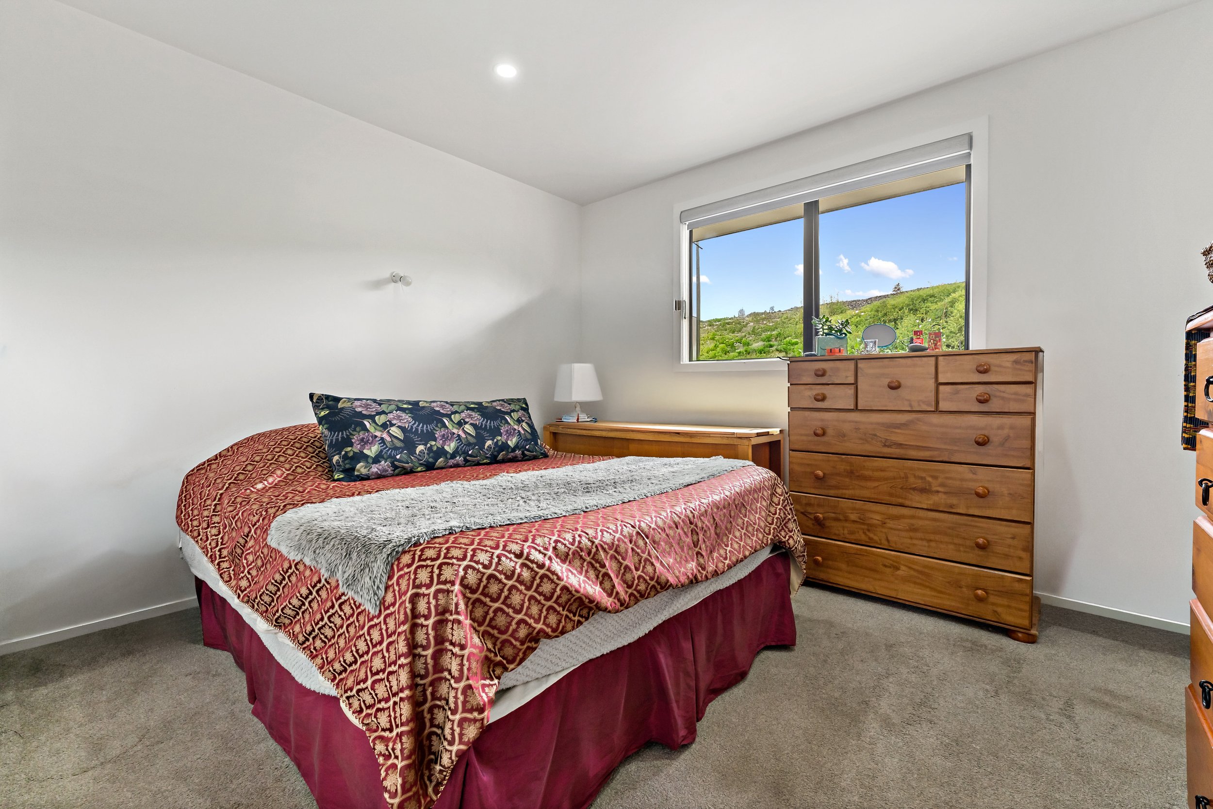A bedroom with a bed, a wooden dresser, and a window showing a green scenic view.