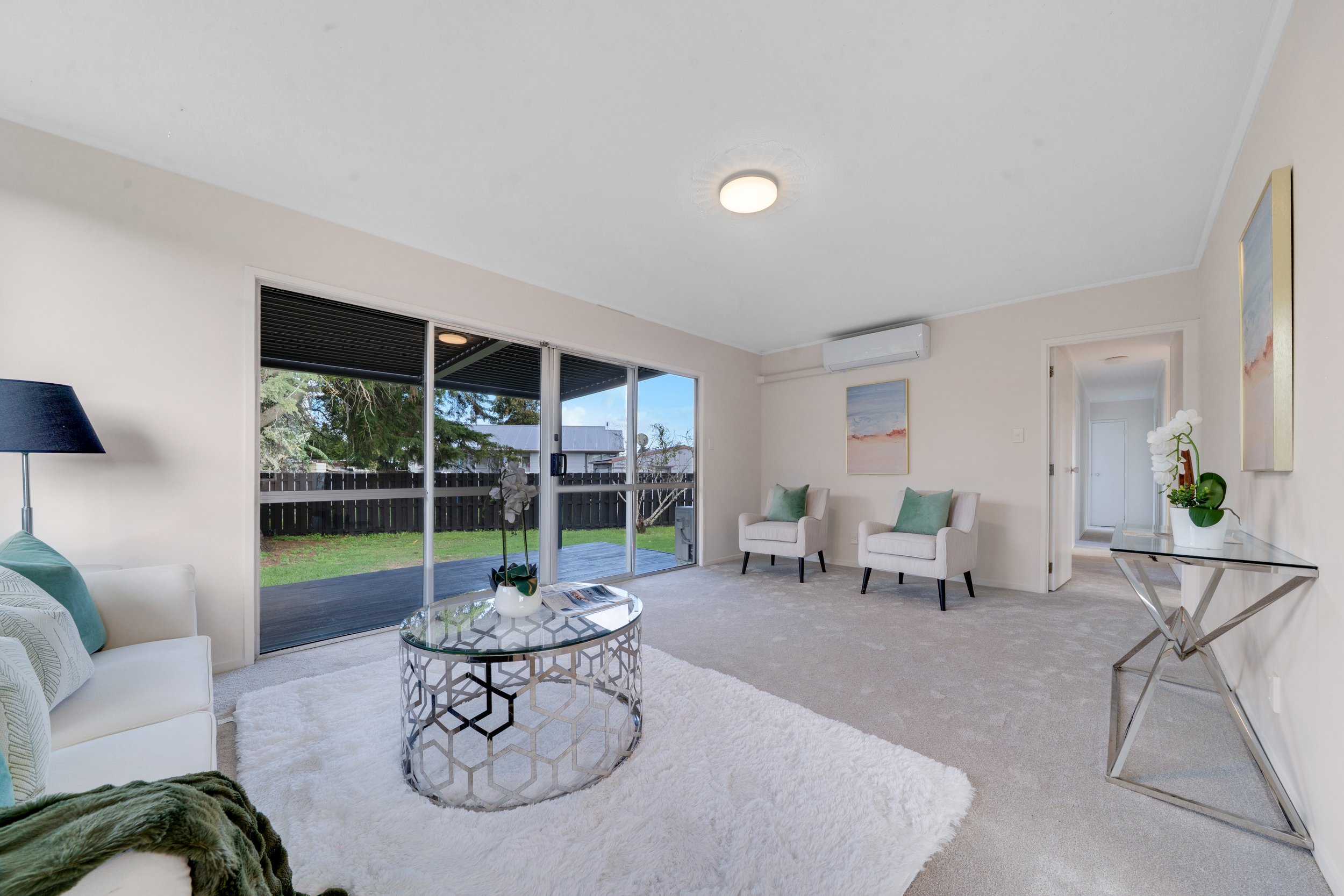 Living room with sliding glass door leading to backyard, white sofa with green pillows, round glass coffee table with decorative metal base, two cream armchairs with green pillows, abstract landscape art on white walls, silver console table with whit