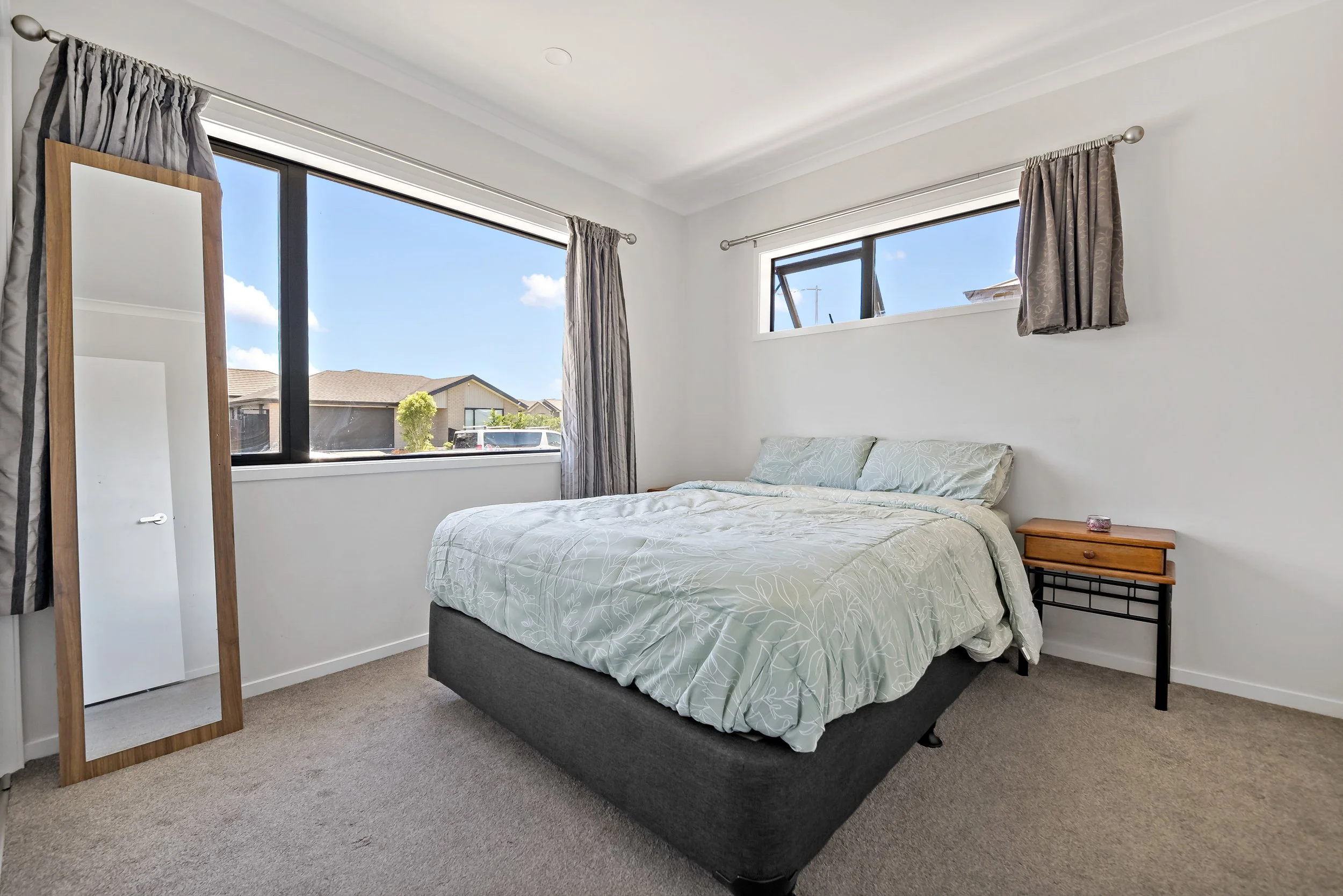 A bedroom with a bed, nightstand, and mirror. Large windows with curtains let in natural light. White walls and beige carpet.