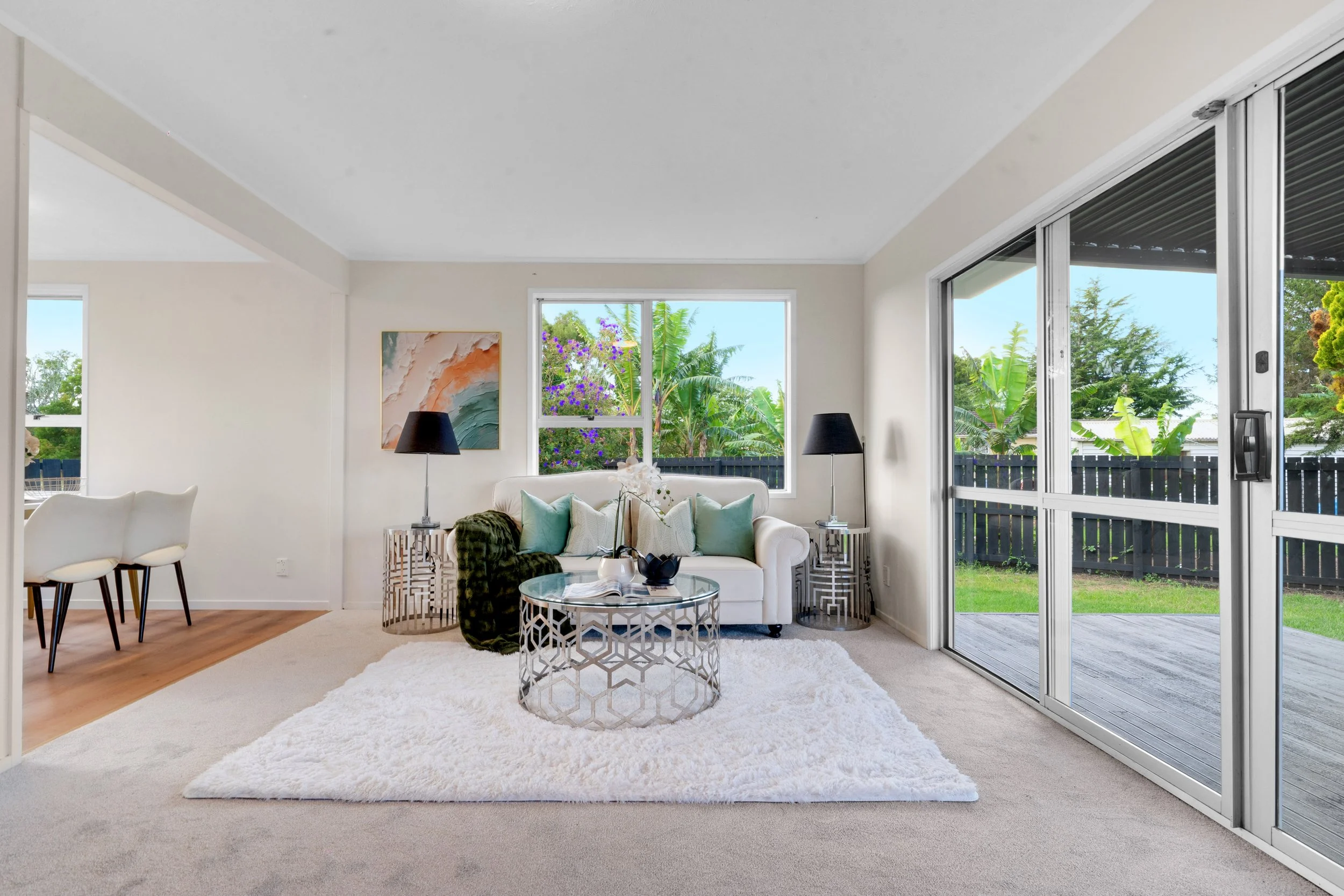 Bright living room with white sofa, green and white throw pillows, black table lamps, glass coffee table, white shag rug, large sliding glass doors leading to backyard, window with garden view, abstract wall art, and dining area with white chairs.