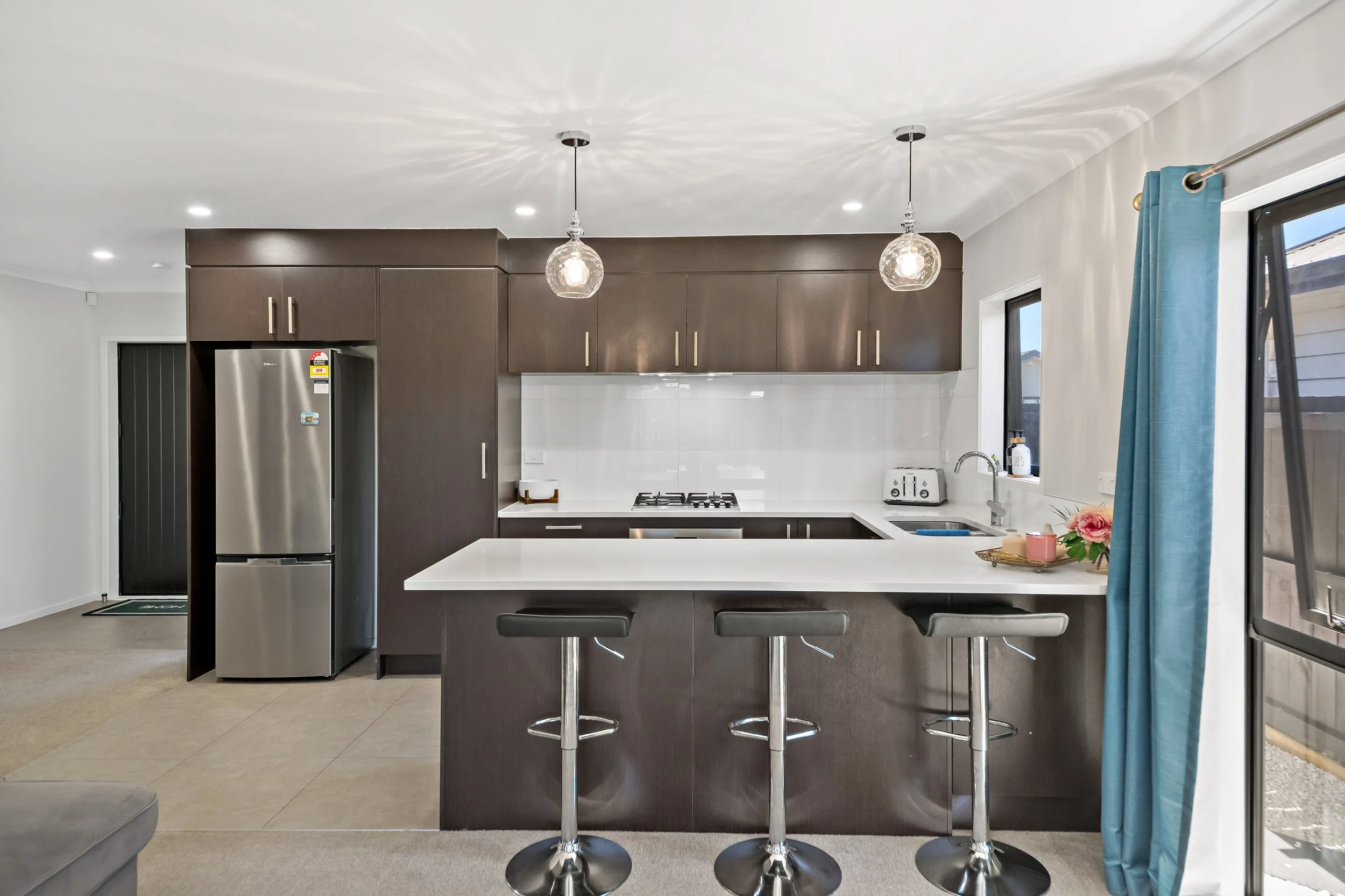 Modern kitchen with dark wood cabinets, white countertops, stainless steel refrigerator, stovetop, toaster, sink, and three bar stools at the island. Light fixtures hang from the ceiling, and there are windows with blue curtains.