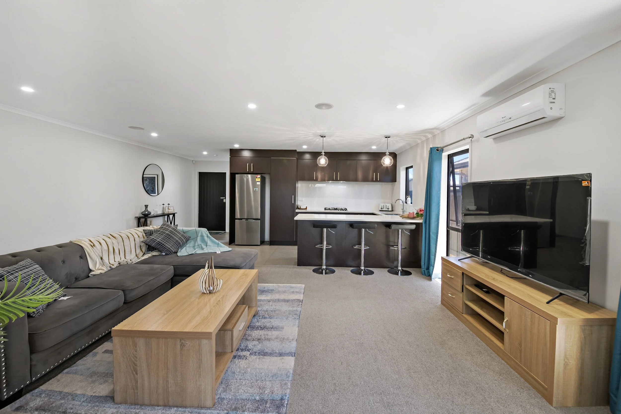 Spacious open-plan living room and kitchen with a gray sectional sofa, wooden coffee table, flat-screen TV on a matching wooden stand, black kitchen cabinetry, stainless steel refrigerator, white countertops, bar stools, and large windows with blue c