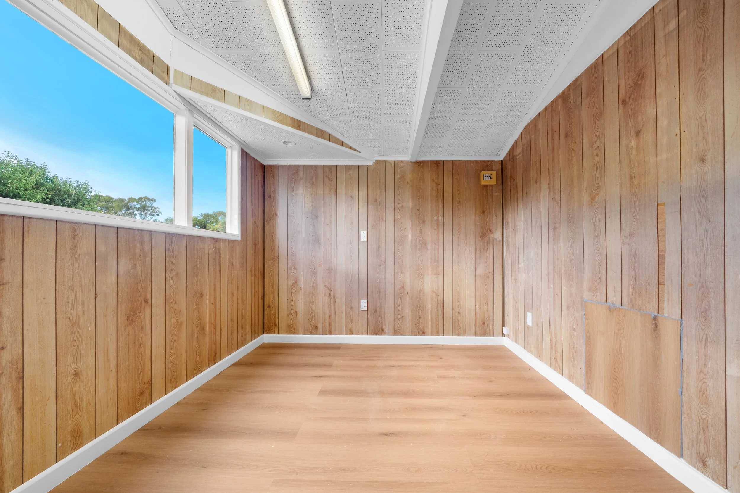 Empty room with wood-paneled walls, a large window showing greenery outside, a white baseboard, and light wood flooring.