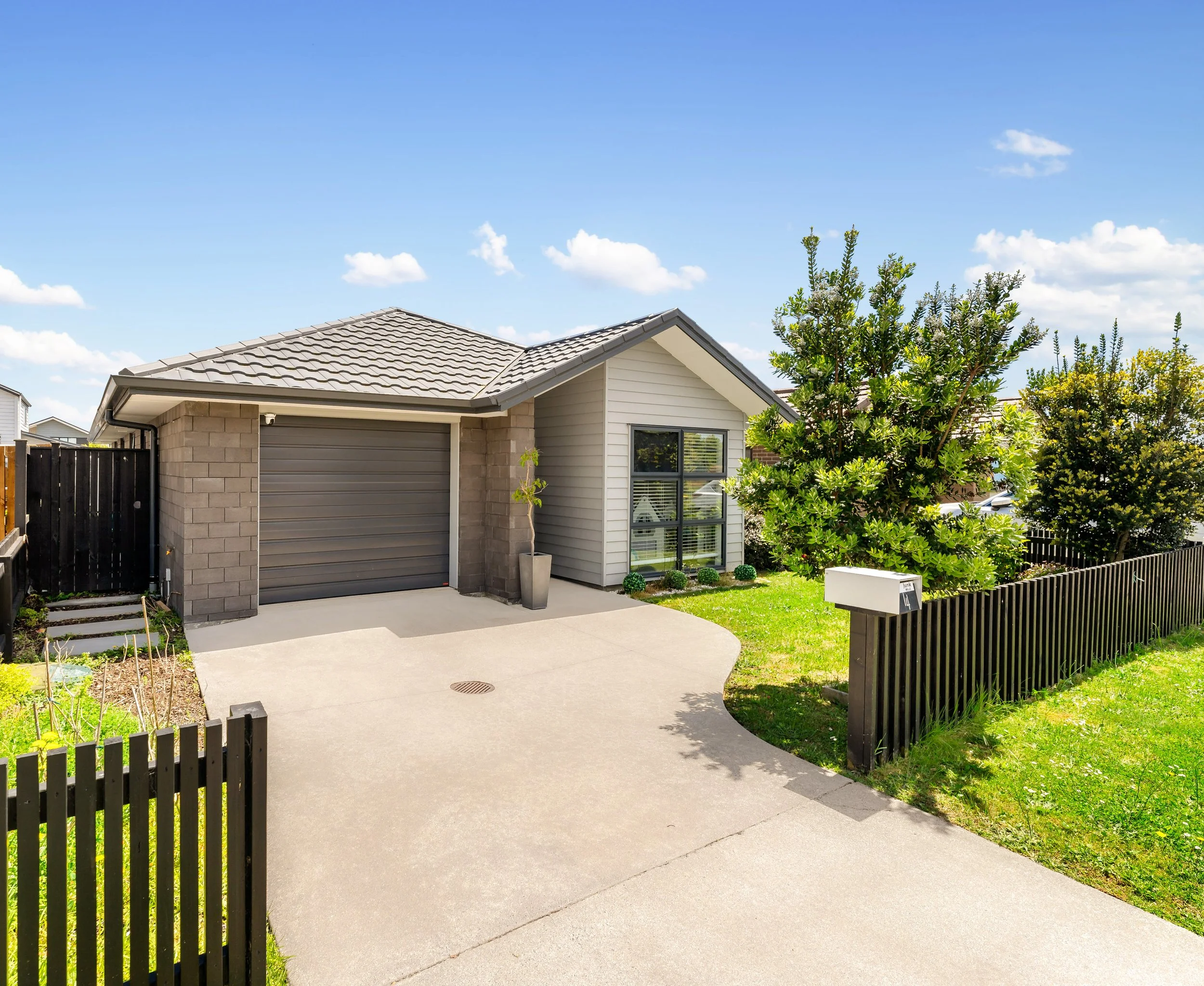 A modern single-story house with a grey tiled roof, beige brick walls, and white siding. It has a closed grey garage door, a small driveway with a drain, and a black wooden fence. There are trees and shrubs in the front yard, a mailbox, and a clear blue sky with a few clouds.