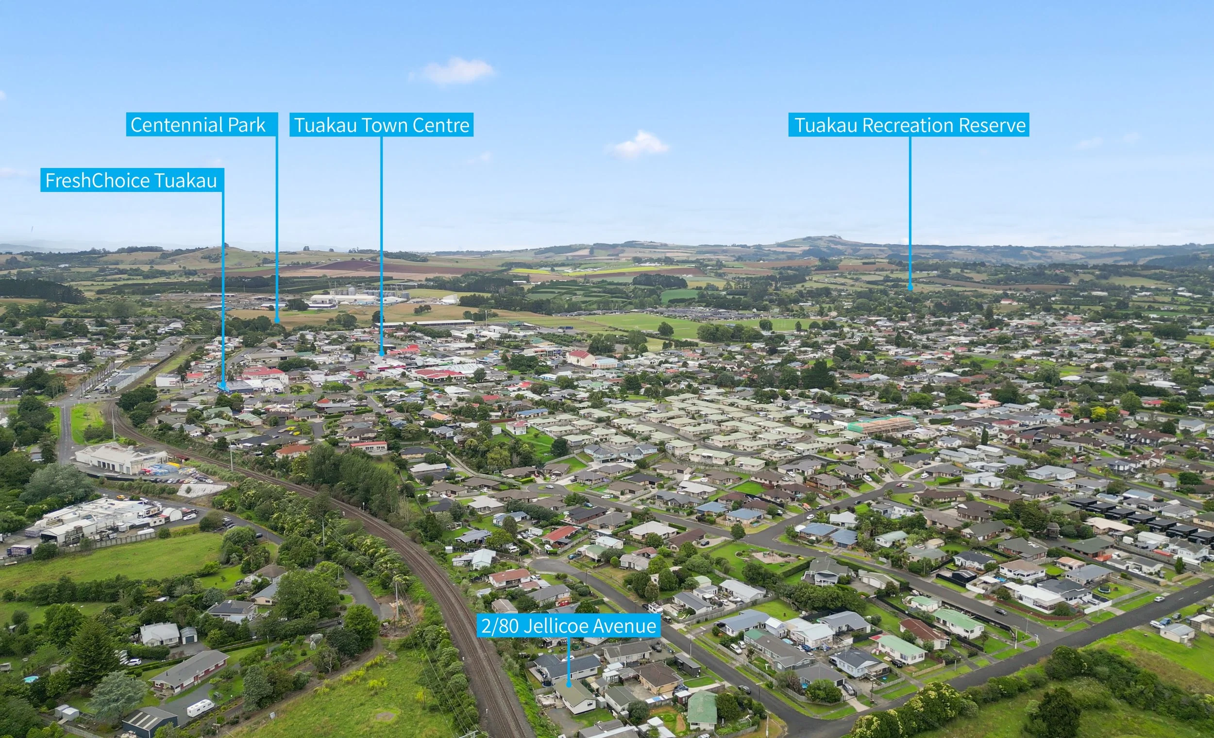 Aerial view of a suburban town with labels indicating locations such as Centennial Park, Tuakau Town Centre, FreshChoice Tuakau, Tuakau Recreation Reserve, and 2/80 Jellicoe Avenue, with green hills and fields in the background.