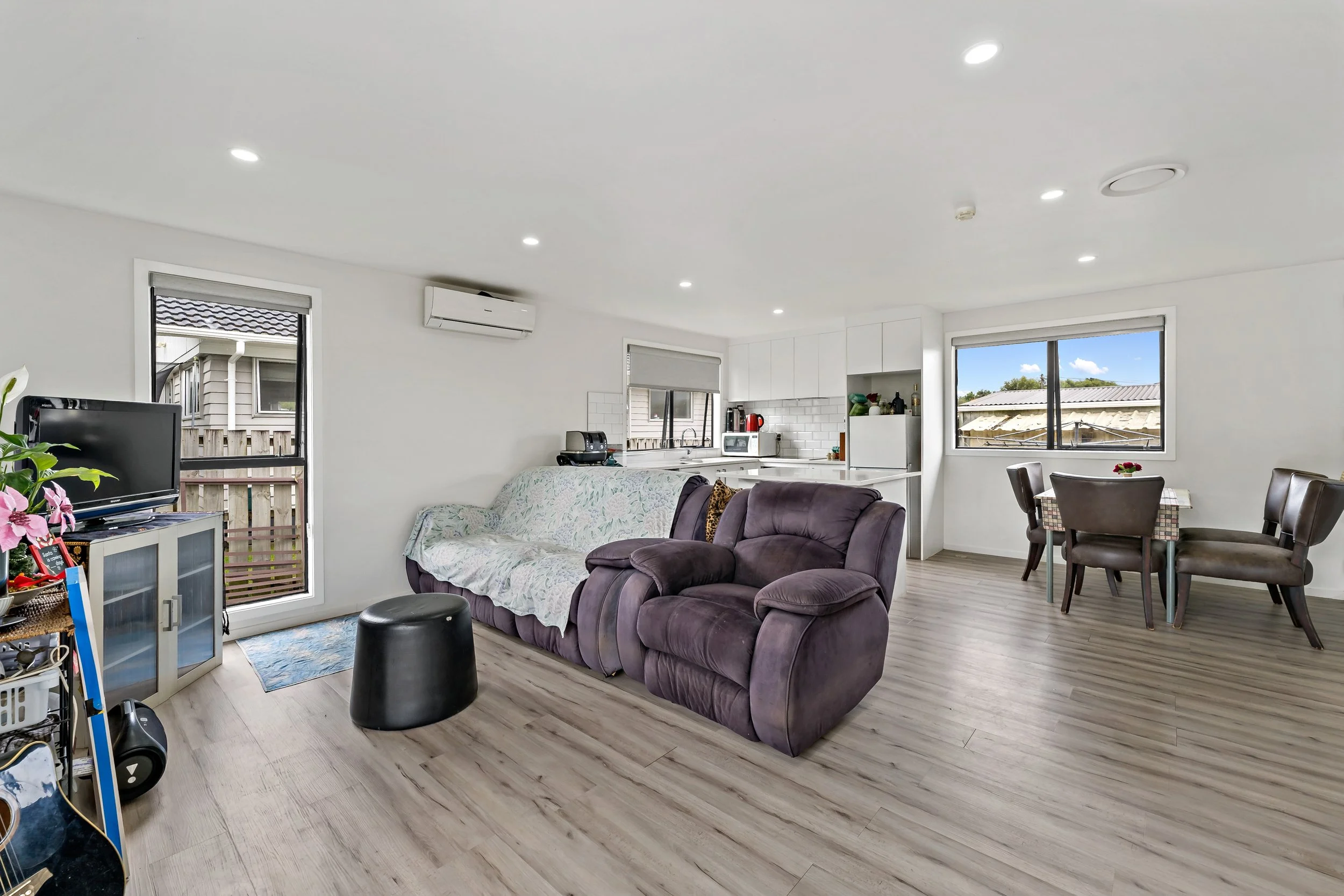 Open living space with purple sofa, small black stool, TV on a stand, dining table with four chairs, and kitchen with white cabinets and appliances. Large windows showing outdoors and an air conditioning unit on the wall.
