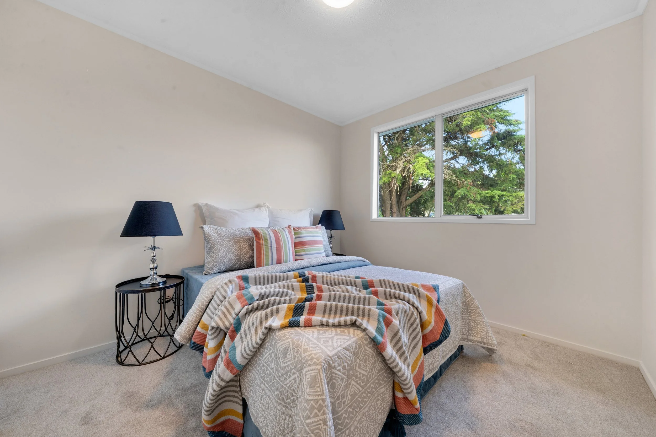 A bedroom with a bed, striped blanket, and pillows near a window showing trees outside.