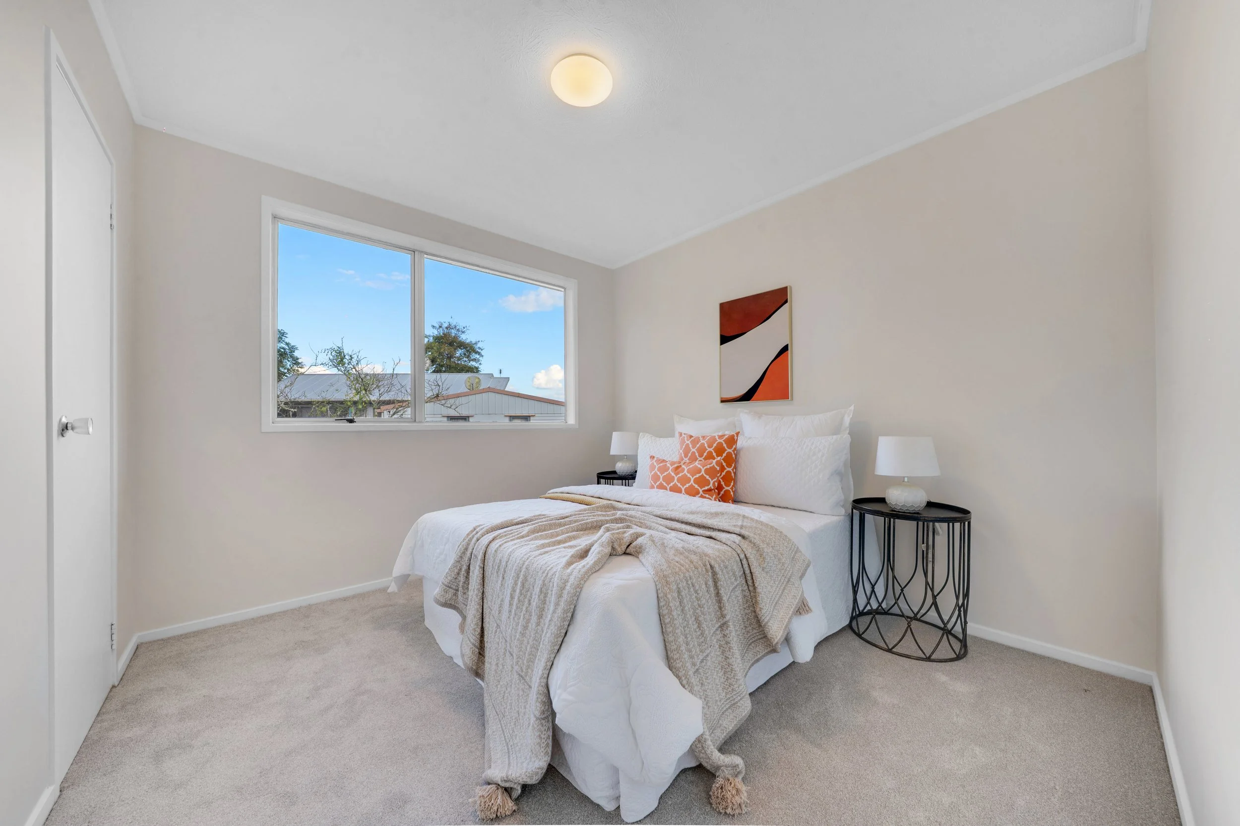 A bedroom with a white bed, orange decorative pillows, beige blanket, two black bedside tables with lamps, a window showing trees and a building outside, and a framed abstract art piece on the wall.