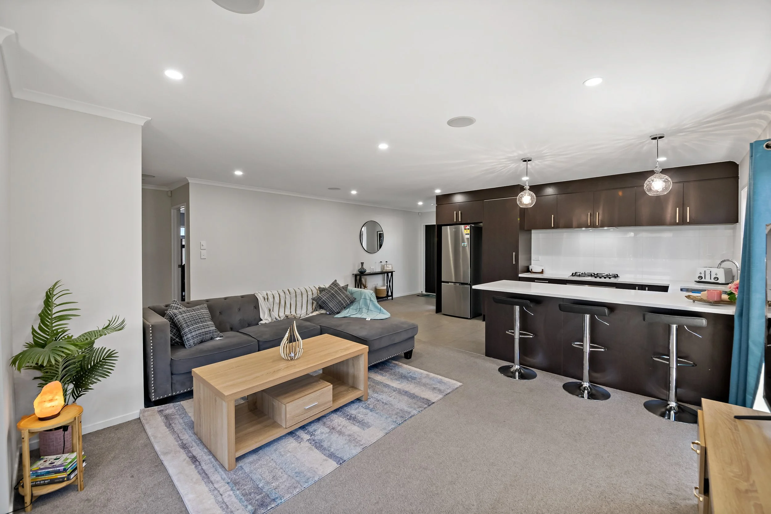 Open-concept living room with gray sectional sofa, light wood coffee table, and bar stools at kitchen island with dark cabinetry and white countertops. Kitchen features stainless steel refrigerator and white tiled backsplash, with pendant lights hang