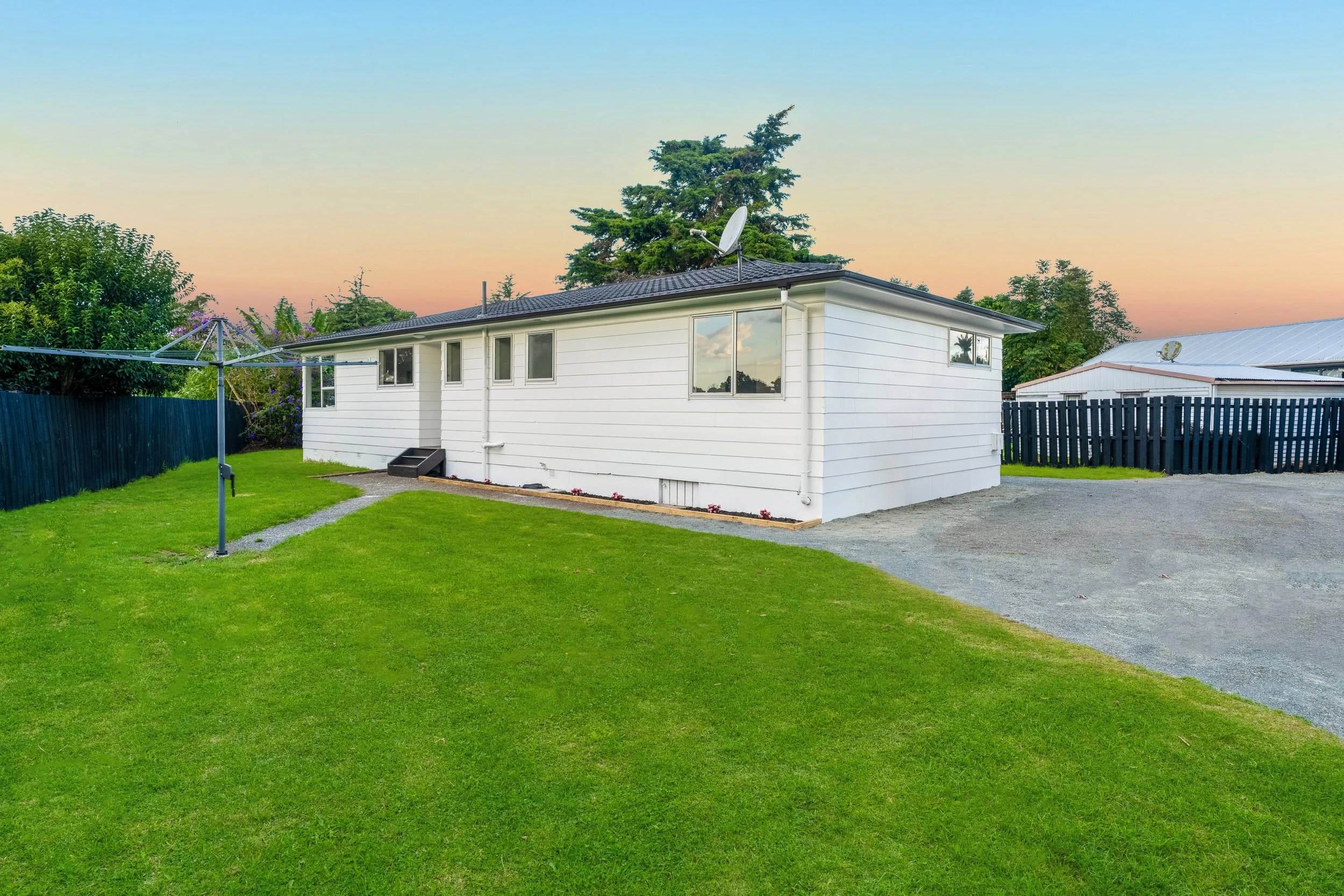 Backyard with a white house, green lawn, gravel driveway, and a rotary clothesline, during sunset.