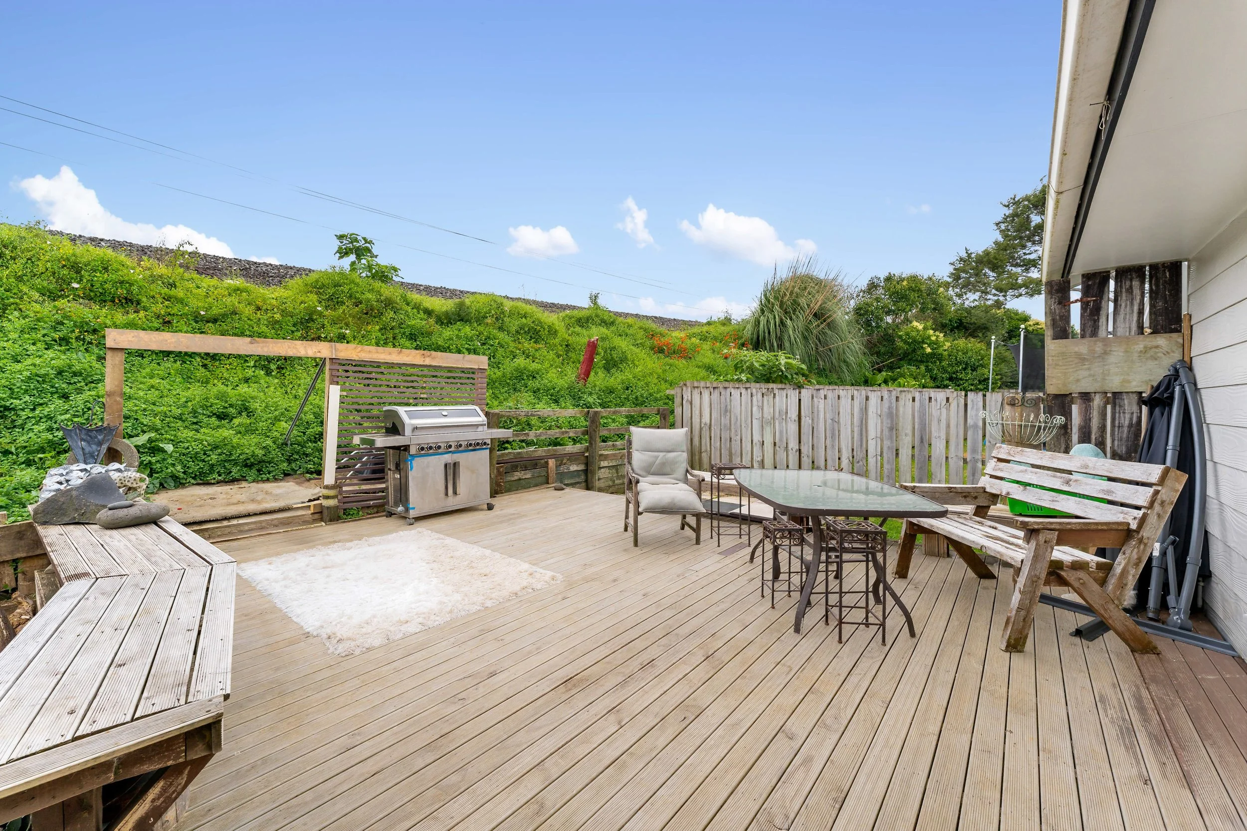 Wooden deck outdoor patio with patio furniture, a small grill, and surrounding greenery, with a fence and hill in background.