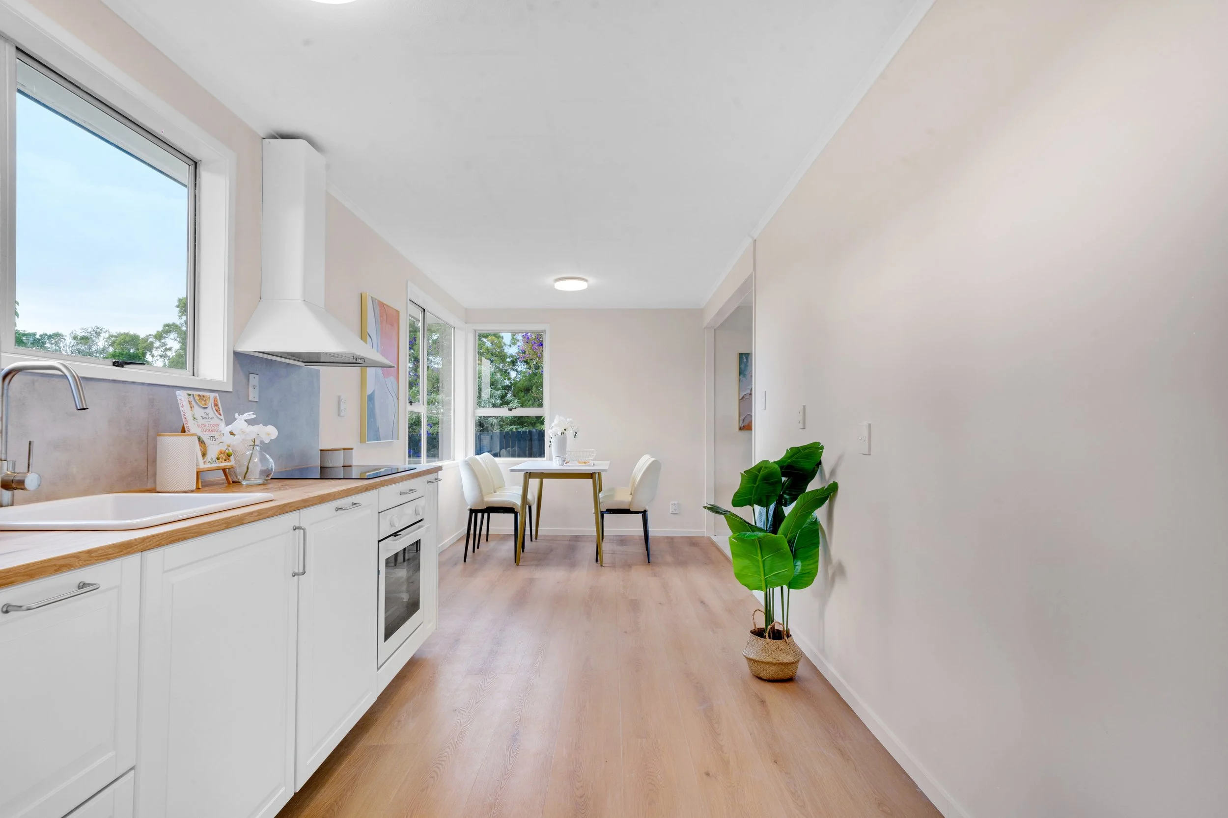 Bright kitchen and dining area with white cabinets, wooden countertop, a plant in a woven basket, and a small dining table with four chairs near large windows with greenery outside.