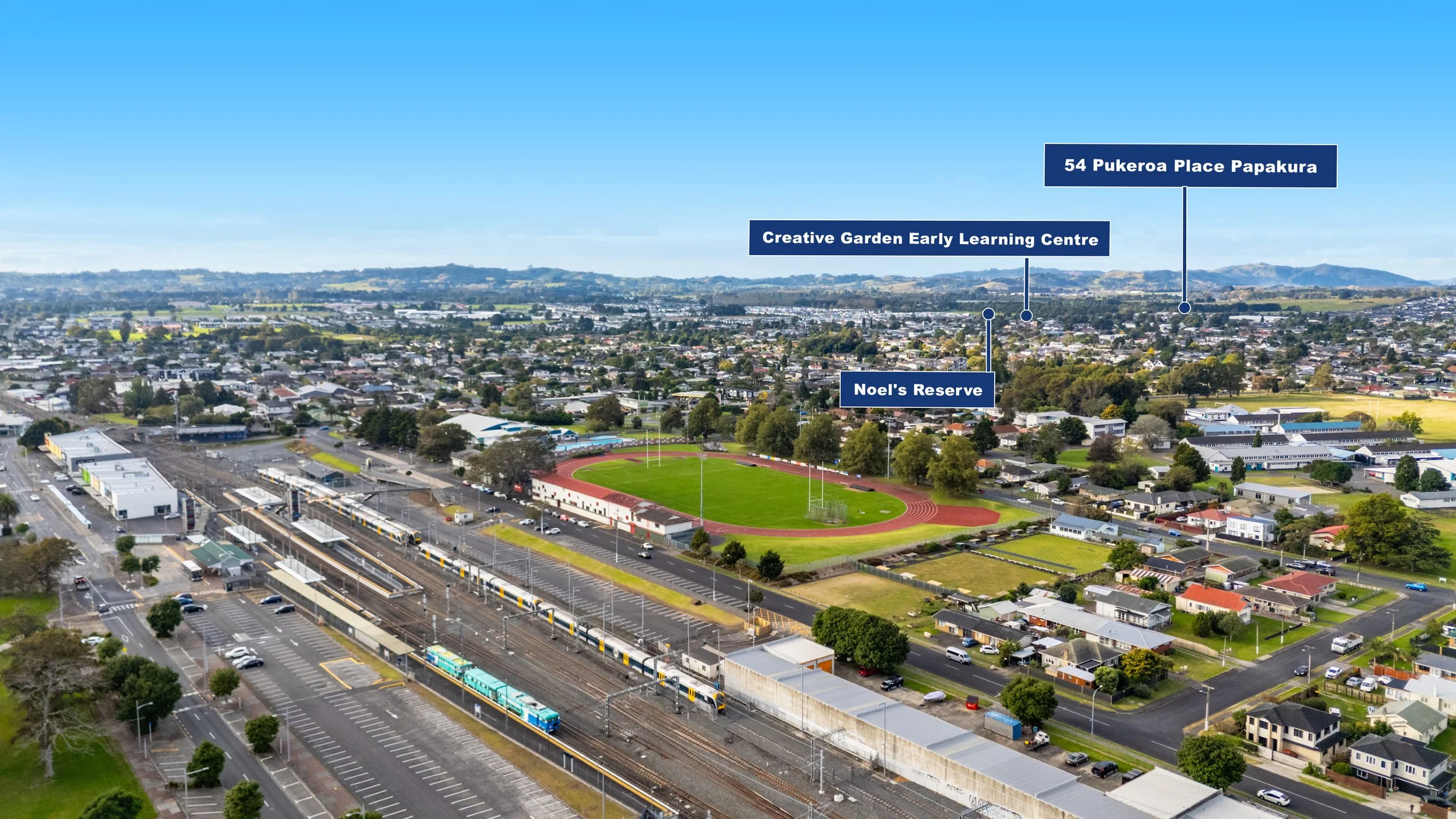 Aerial view of a city neighborhood featuring a sports oval with green field and running track, labeled as Noel's Reserve, surrounded by residential houses, a train station with trains, parking lots, and commercial buildings. Landmarks labeled include