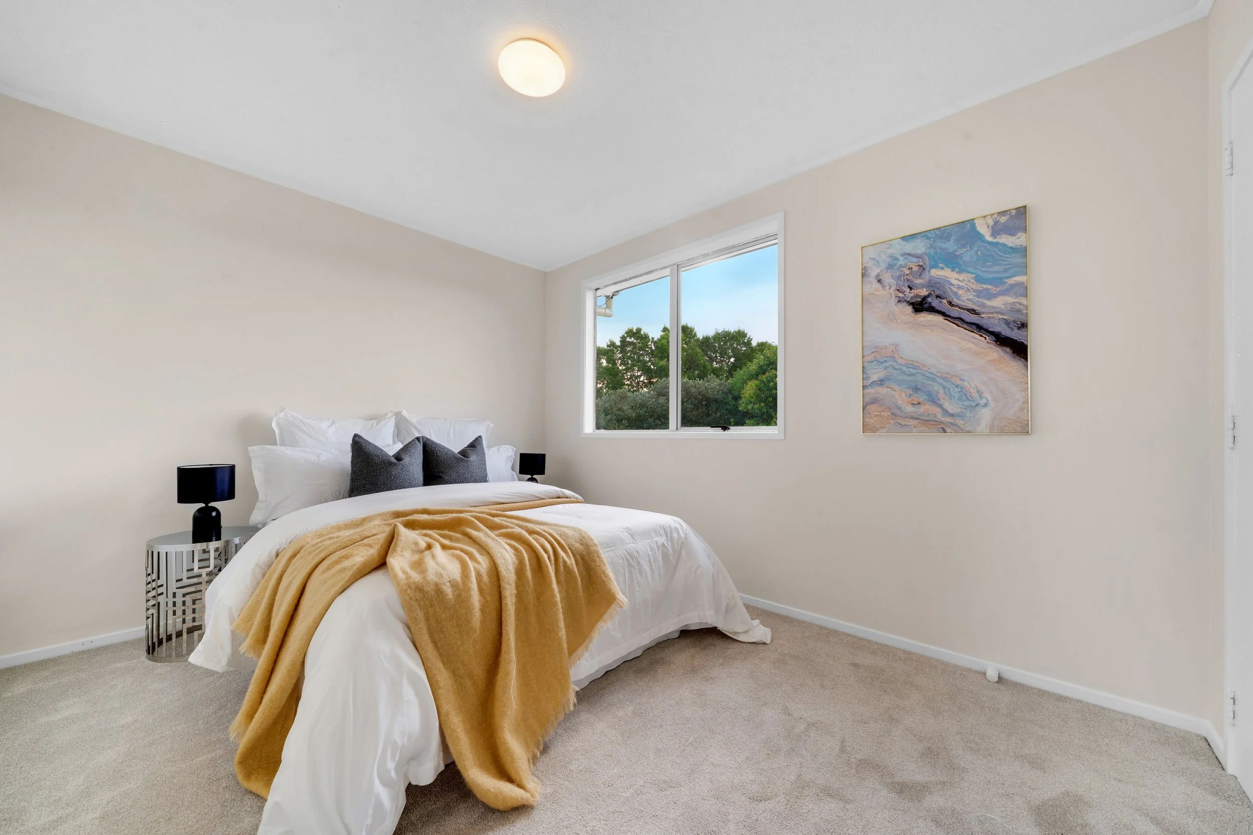 A bedroom with a bed covered in white bedding, two dark gray pillows, a mustard-colored throw blanket, a window showing trees outside, a contemporary beige wall, an abstract painting, and two small black lamps on a modern black and white side table.