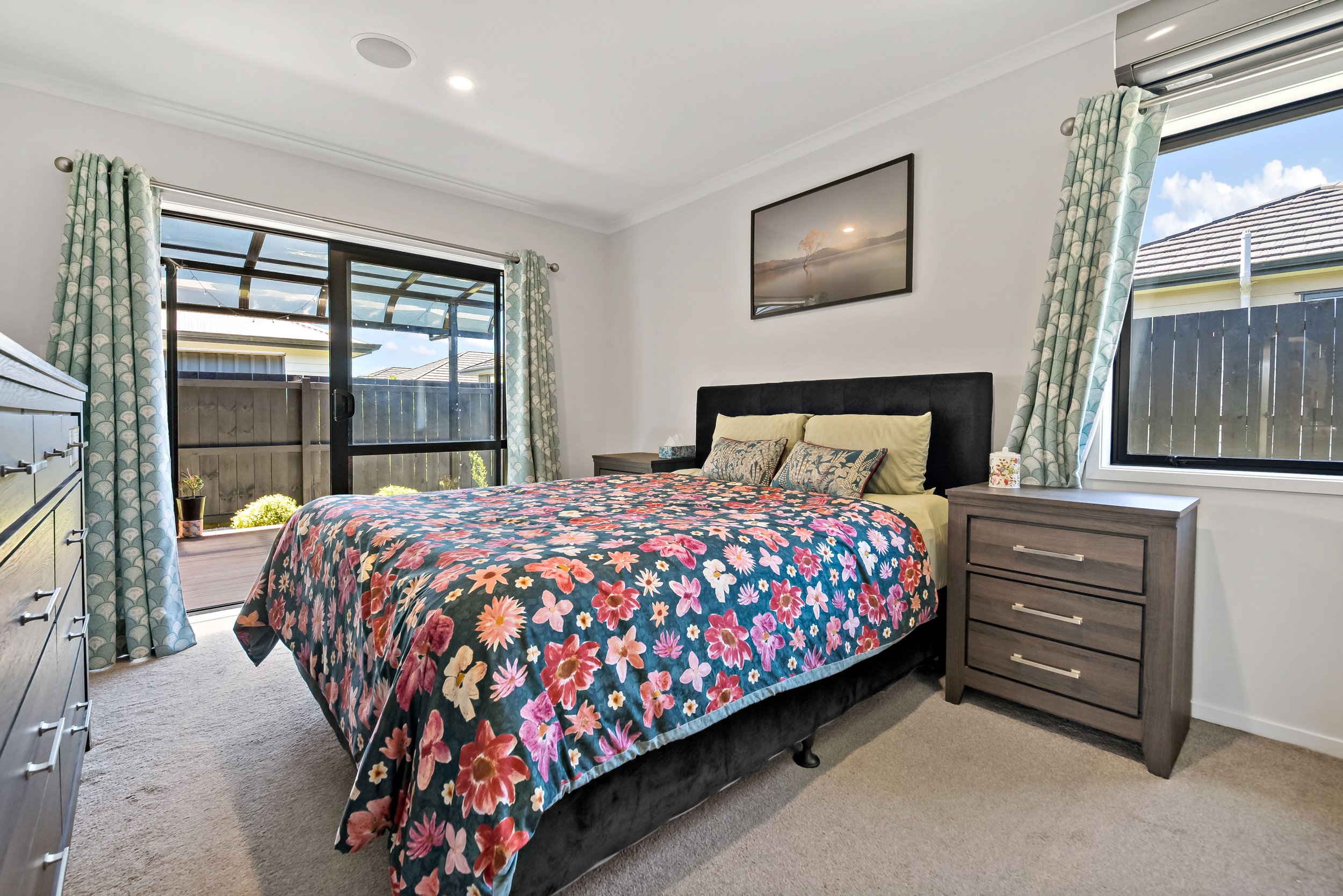 Bedroom with bed, floral quilt, two nightstands, window with curtains, and sliding door leading to outdoor patio.