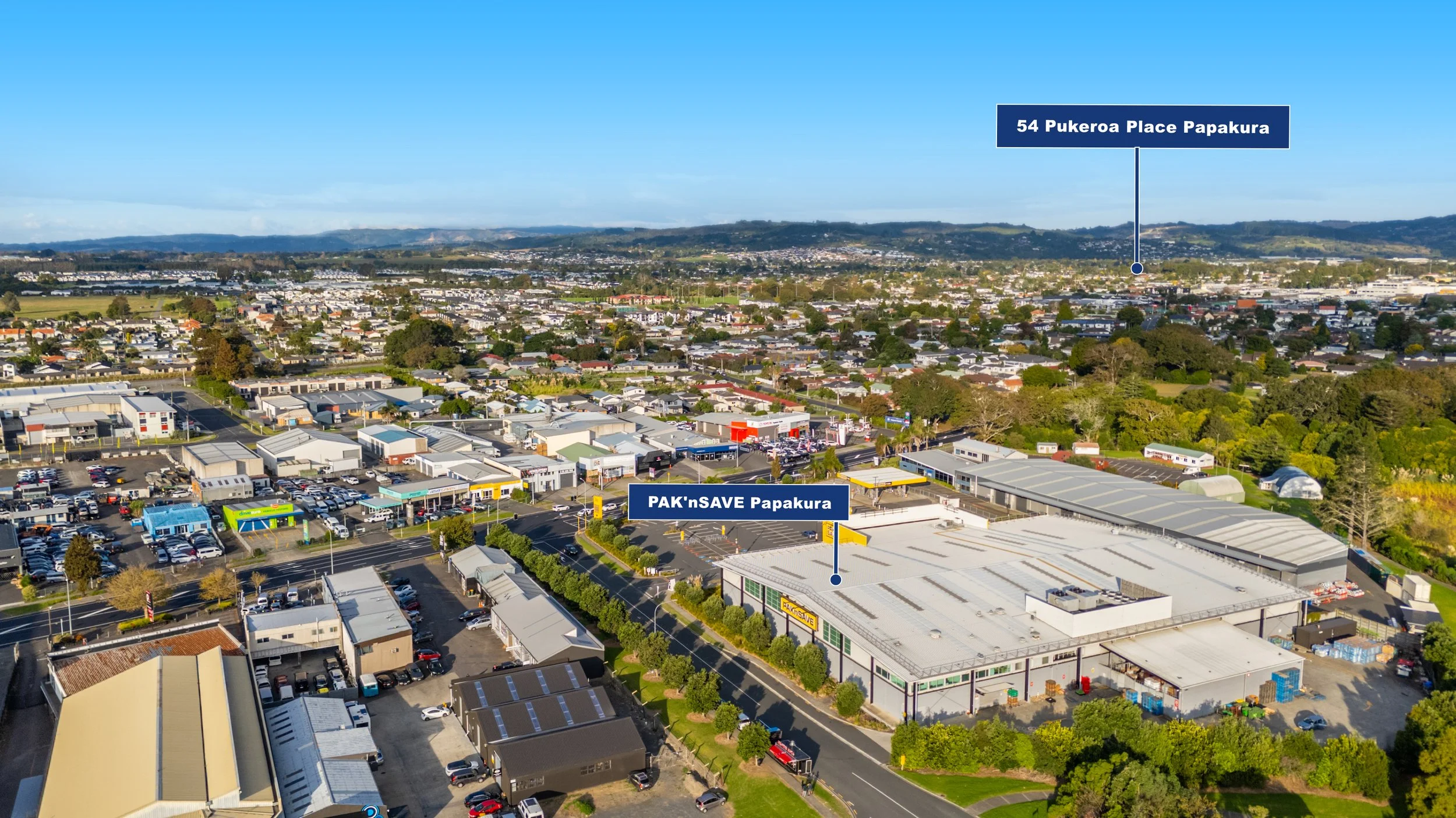 Aerial view of Papakura area with labeled locations: '54 Pukeroa Place Papakura' in the background and 'PAK'nSAVE Papakura' supermarket in the foreground, amidst residential and commercial buildings, greenery, and mountains in the distance.
