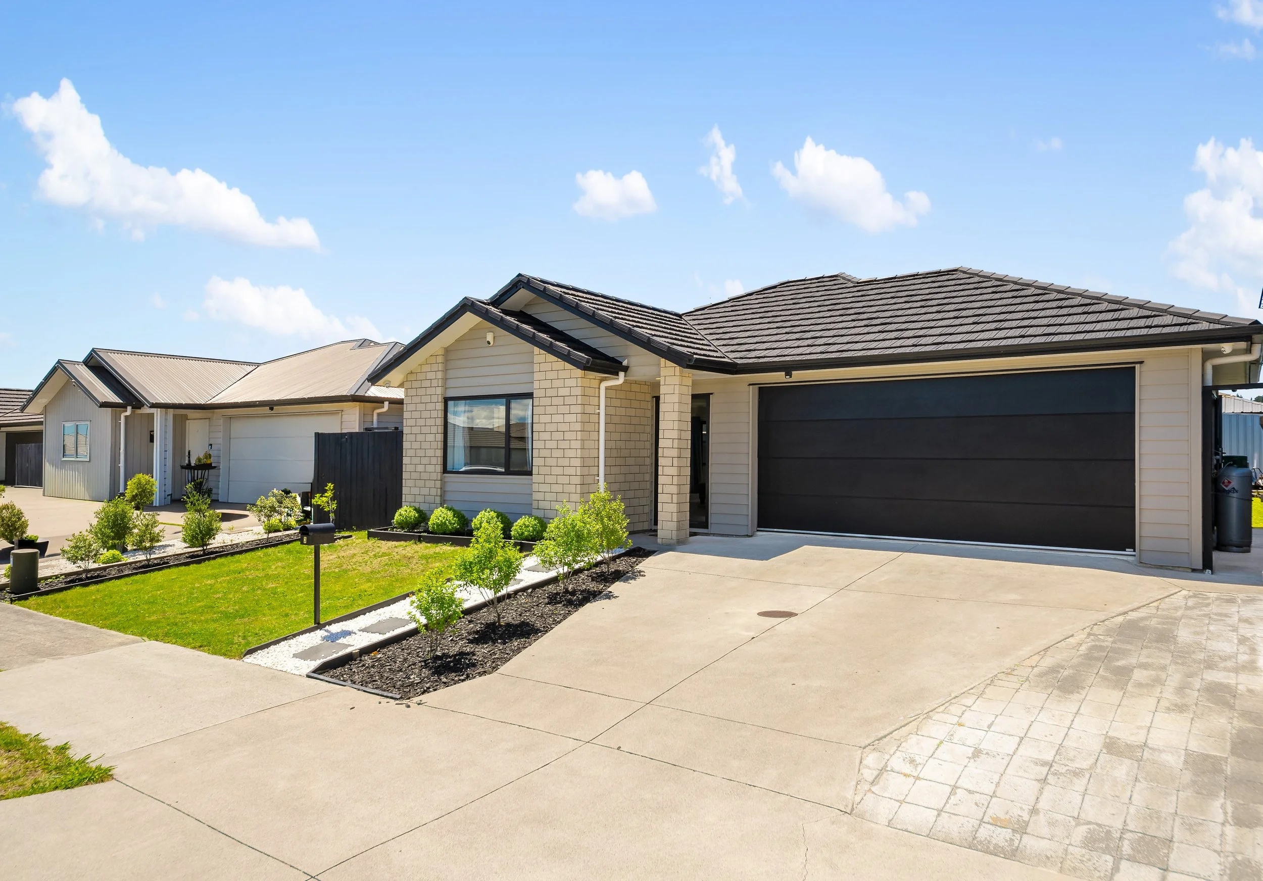 Modern single-story house with a black garage door, brick and siding exterior, manicured lawn, small trees, and a driveway on a sunny day with blue sky and clouds.