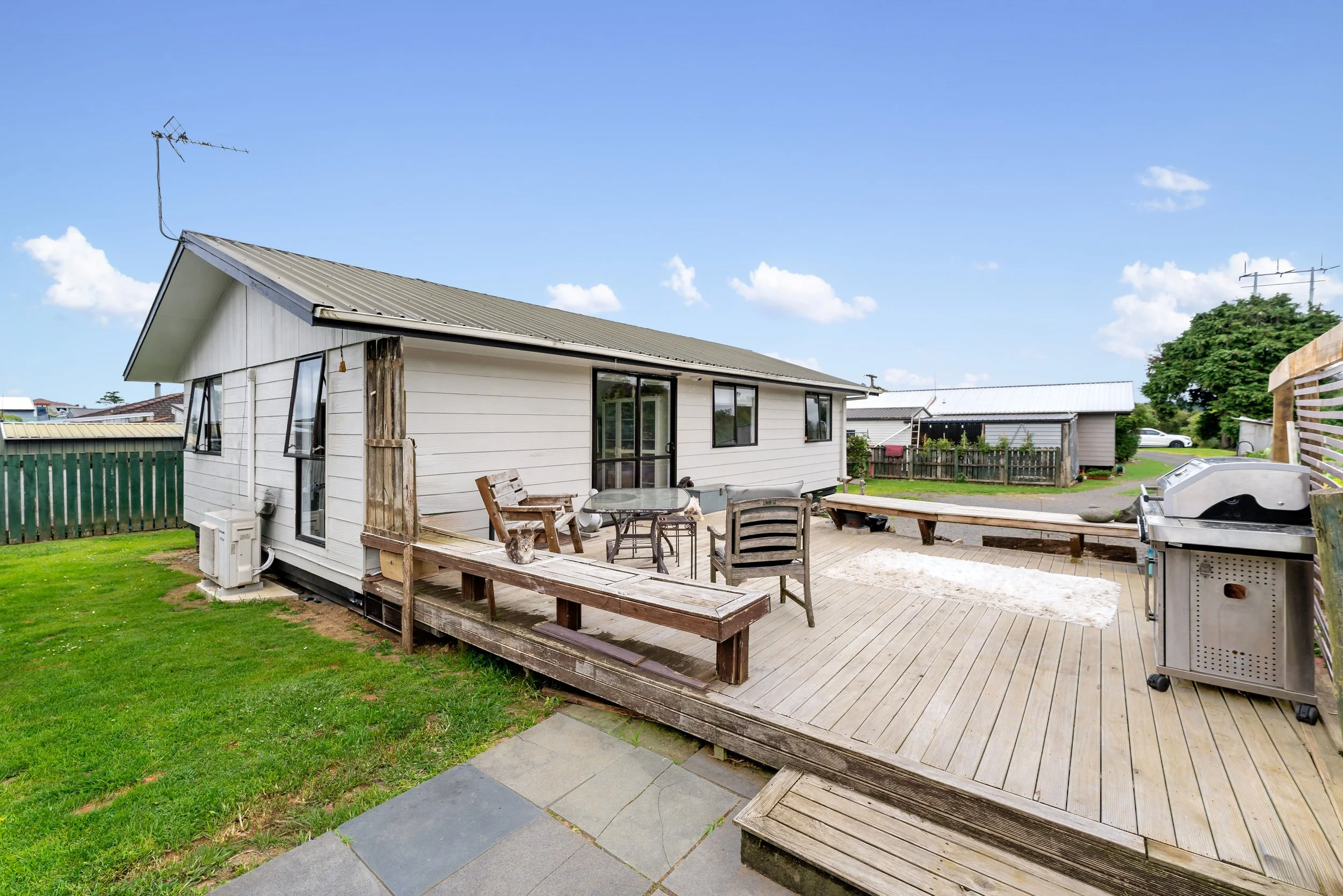 A backyard patio with outdoor furniture, a grill, and a wooden deck attached to a white house with a metal roof, green grass, and neighboring sheds under a blue sky with scattered clouds.