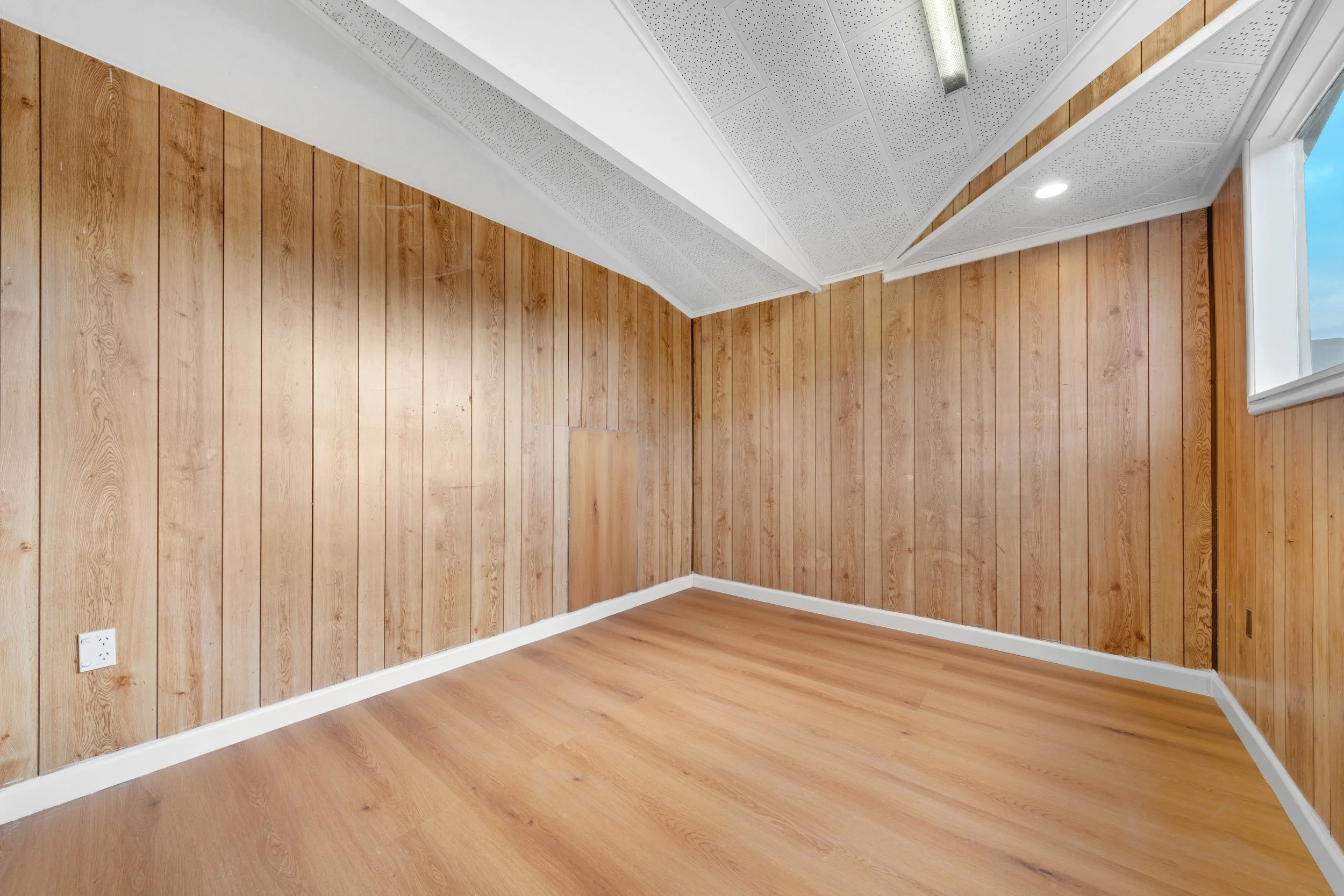 Empty room with wood-paneled walls, a window, and a vinyl floor, with ceiling panels and lighting.