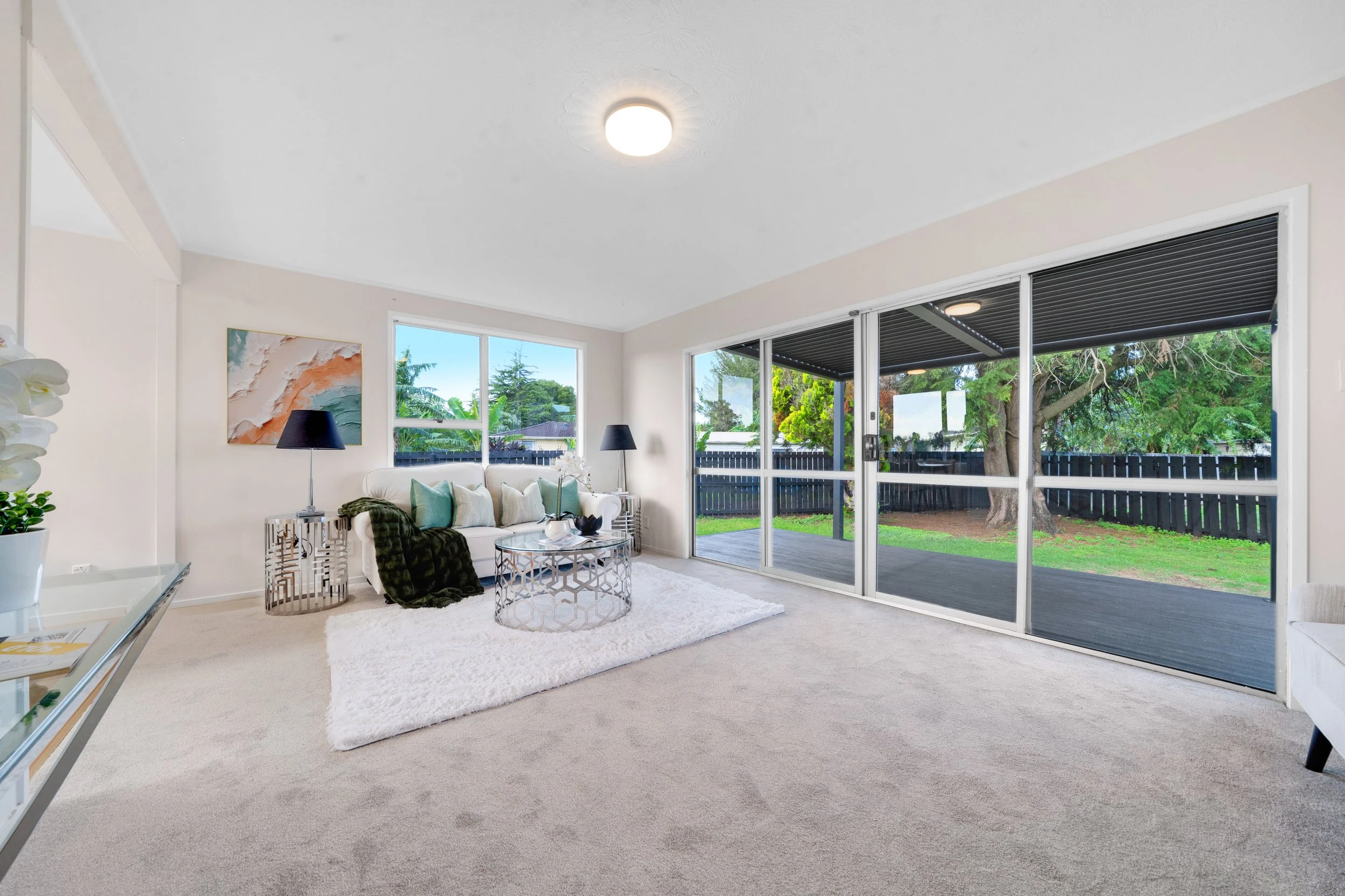 Living room with beige carpet, white sofa with cushions, black lamps, circular coffee table, rug, large sliding glass doors opening to backyard with trees and black fence, and wall art.