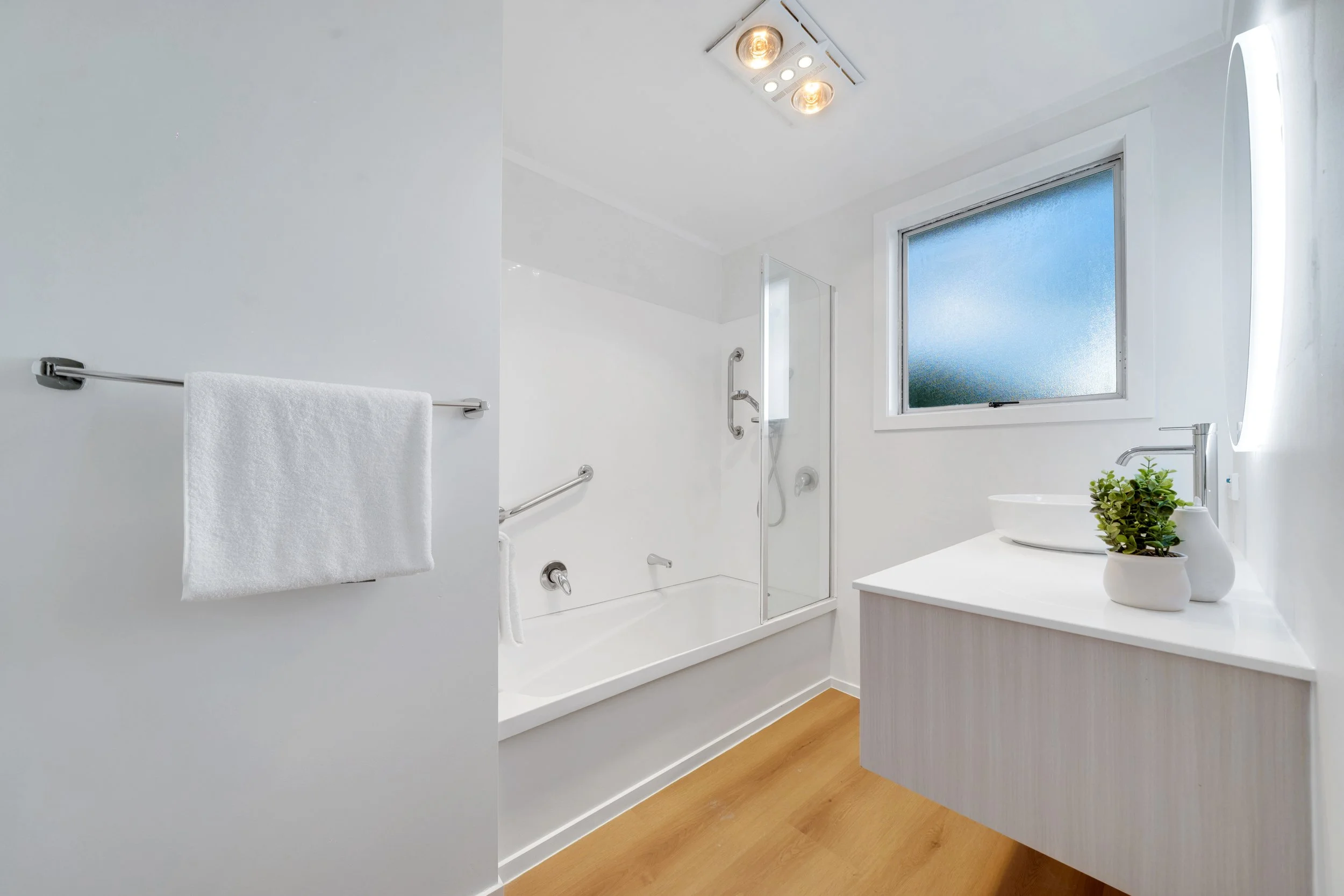 A clean, modern bathroom with a white bathtub, a glass shower door, a window with frosted glass, a white sink with a mirror and a small potted plant, and a white towel hanging on a metal rod.