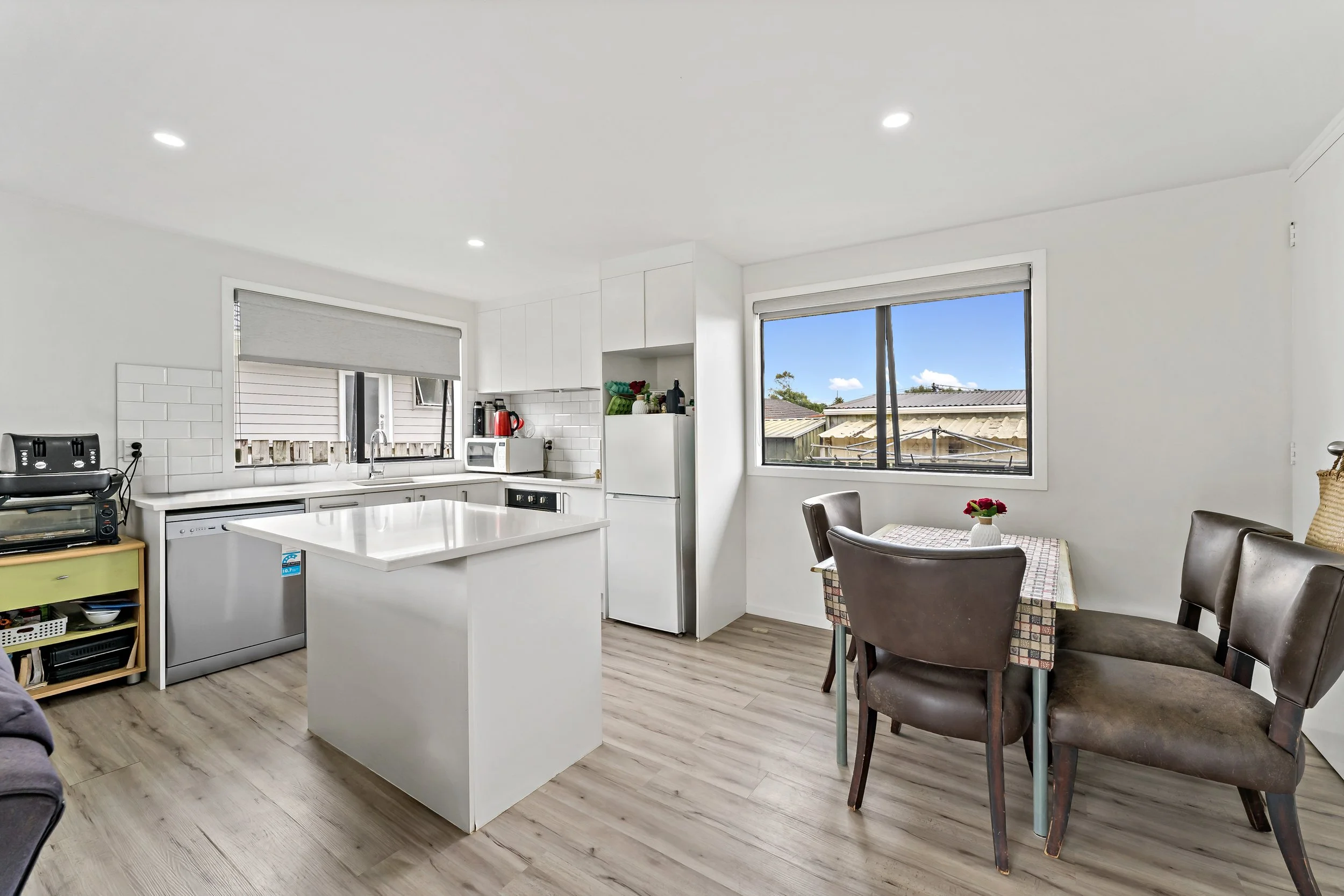 A bright kitchen and dining area with white cabinets, a kitchen island, and a small dining table with four chairs and a vase of flowers, next to large windows showing a blue sky and neighboring rooftops.