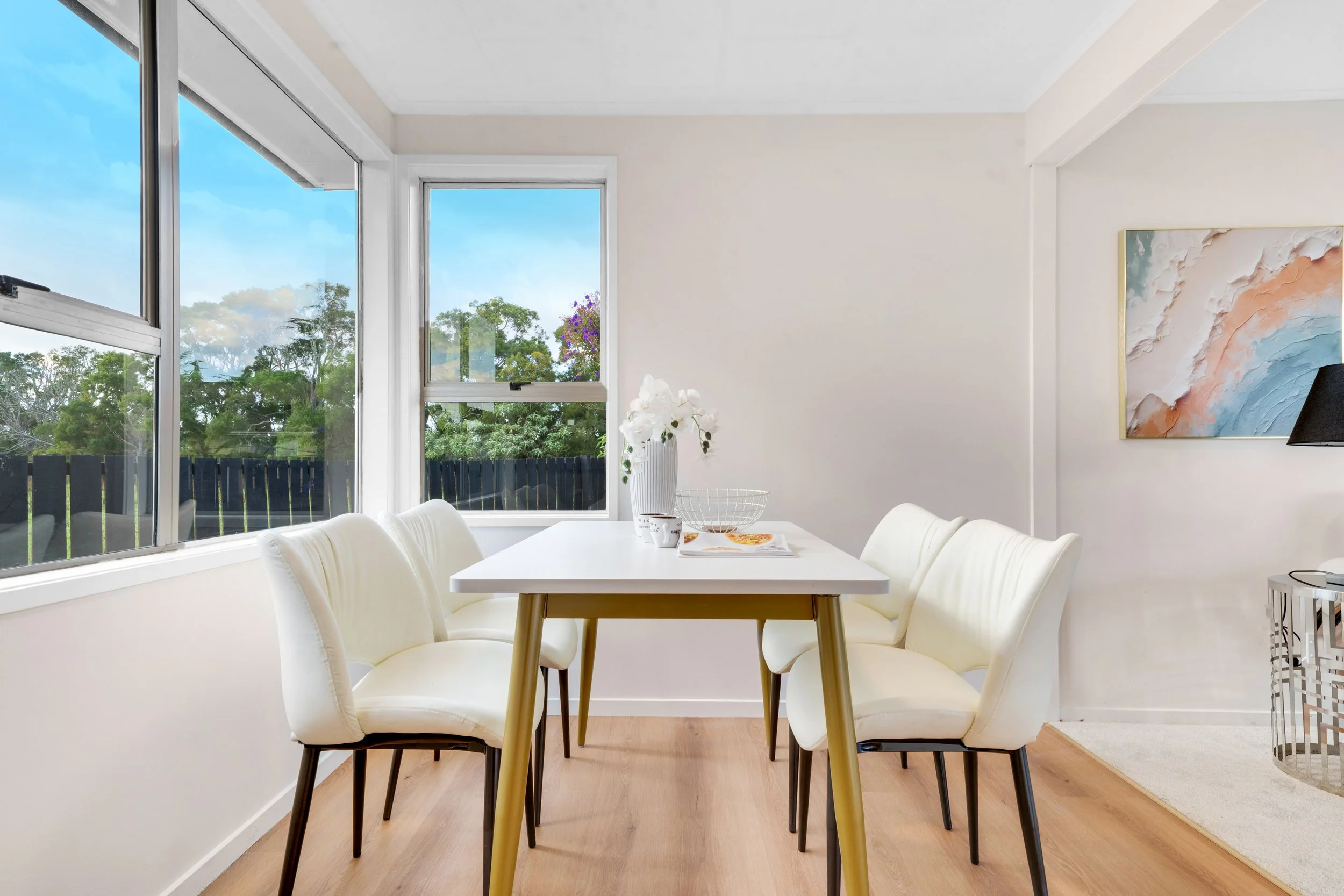 Bright dining room with white table, six white chairs, and large windows showing outdoor trees.