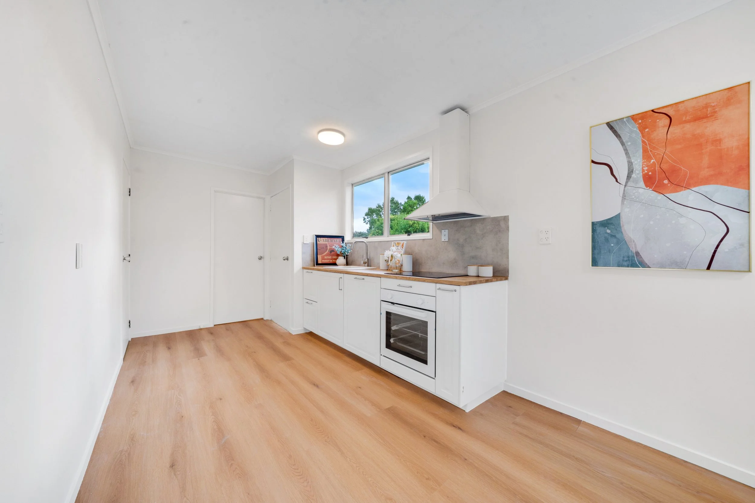Empty kitchen with white cabinets, a window, artwork on the wall, and light wood flooring.
