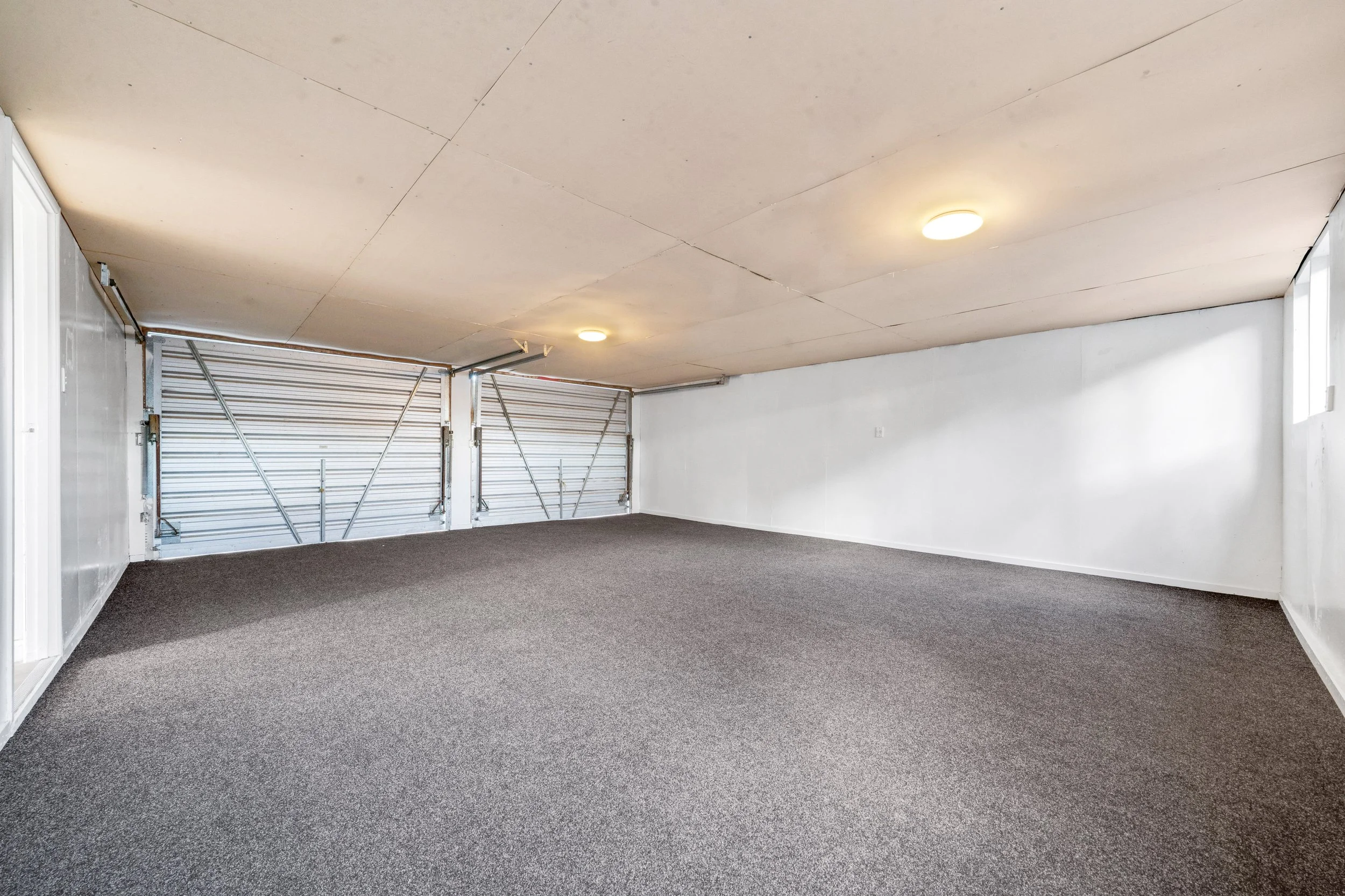 Empty garage with white walls, gray carpeting, and metal garage doors, illuminated by ceiling lights.