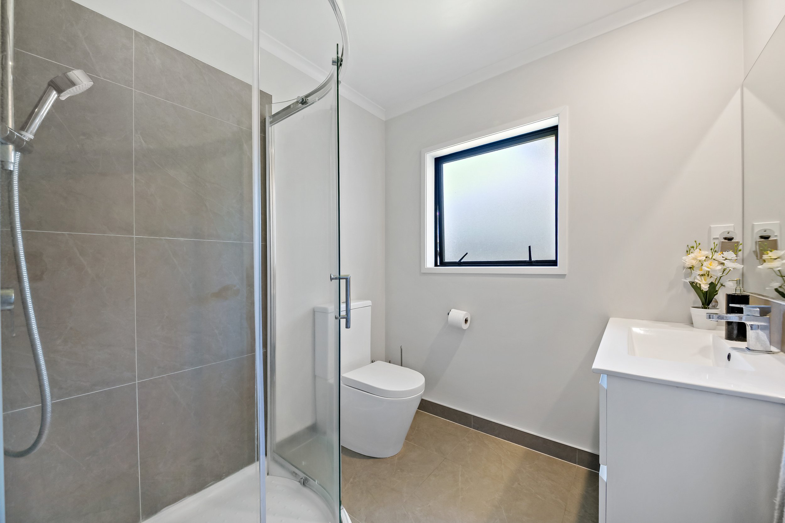 Modern bathroom with a glass shower enclosure, white toilet, window with frosted glass, and a white vanity with a sink and decorative flowers.