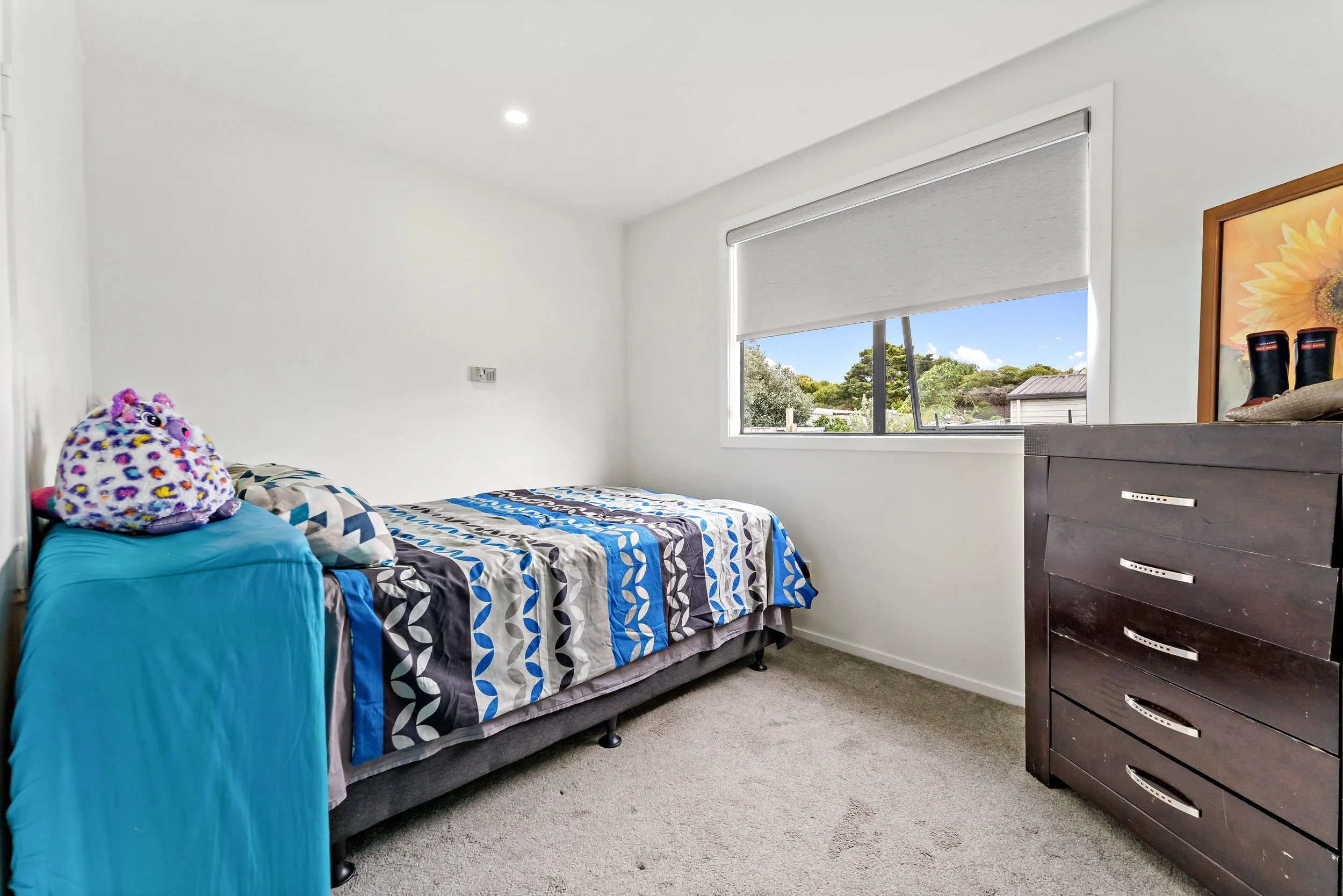 A small bedroom with a bed covered by a patterned quilt, a blue blanket, and a purple plush toy. There is a dark wooden dresser with some items on top, and a window with a roller blind revealing trees and a blue sky outside.