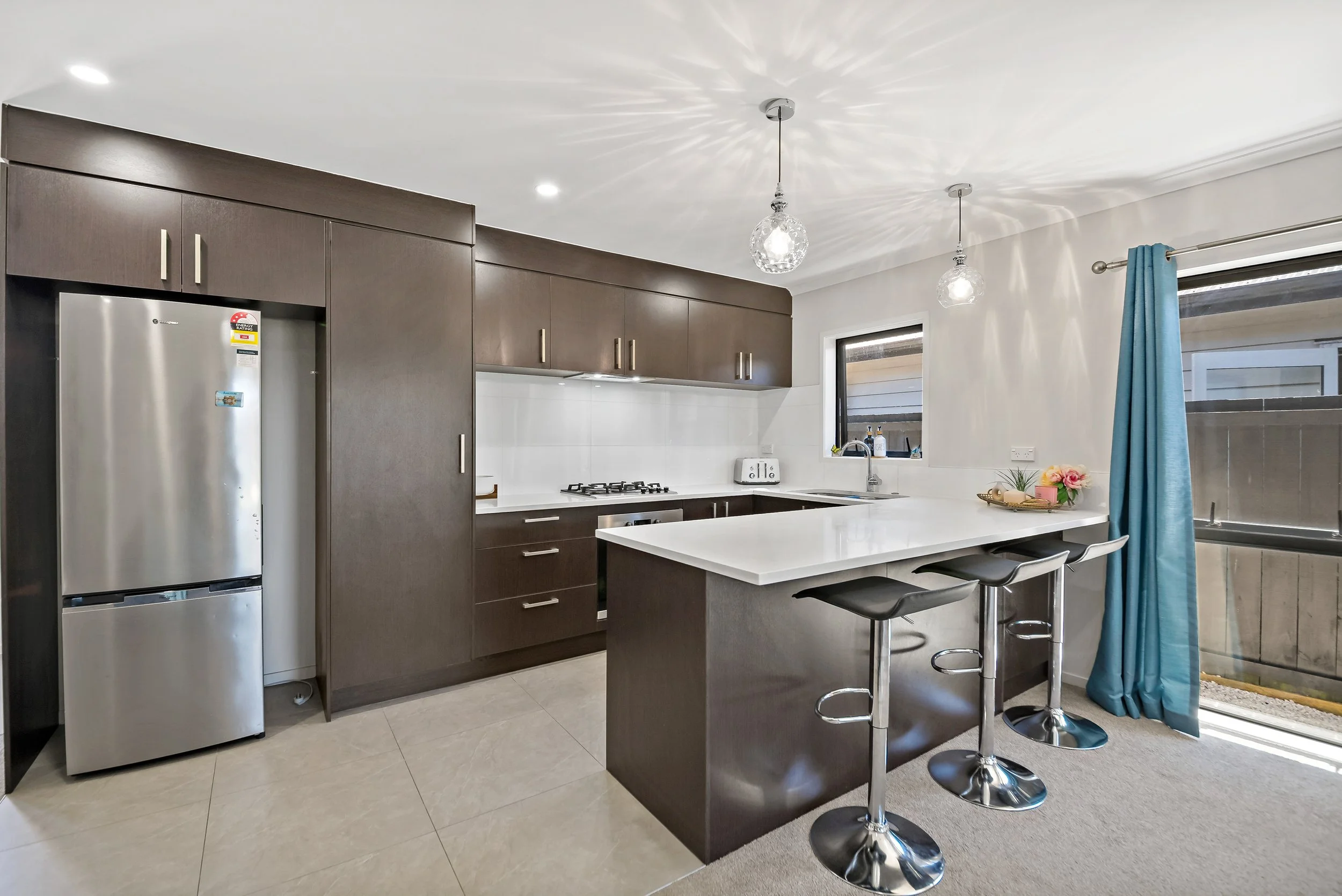 Modern kitchen with dark wood cabinets, white countertop, stainless steel refrigerator, gas stove, white tiled backsplash, two hanging pendant lights, window, and sliding glass door with blue curtains.