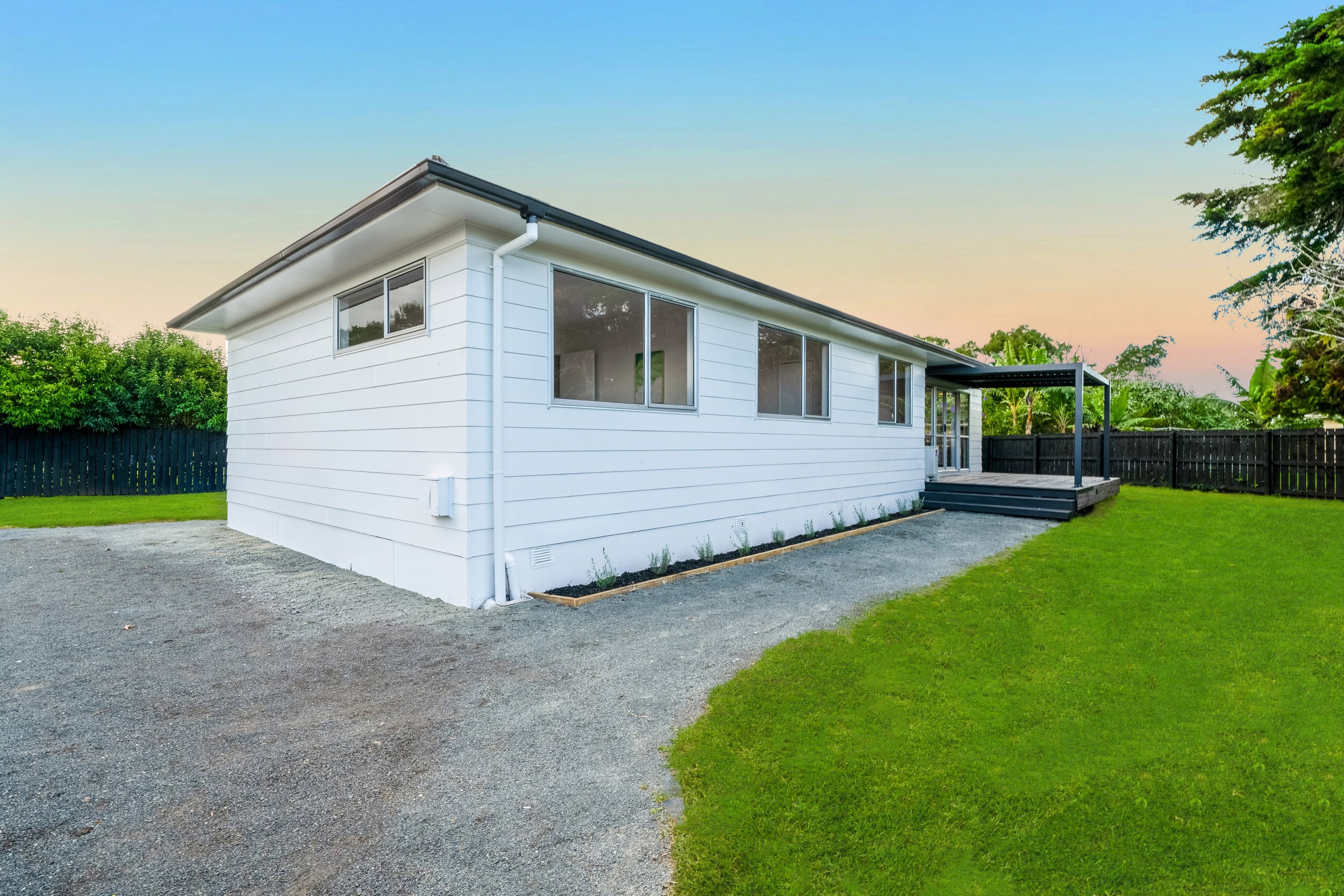 Modern white house with large windows, small deck with stairs, fenced backyard, green grass, gravel driveway, and clear sky at sunset.