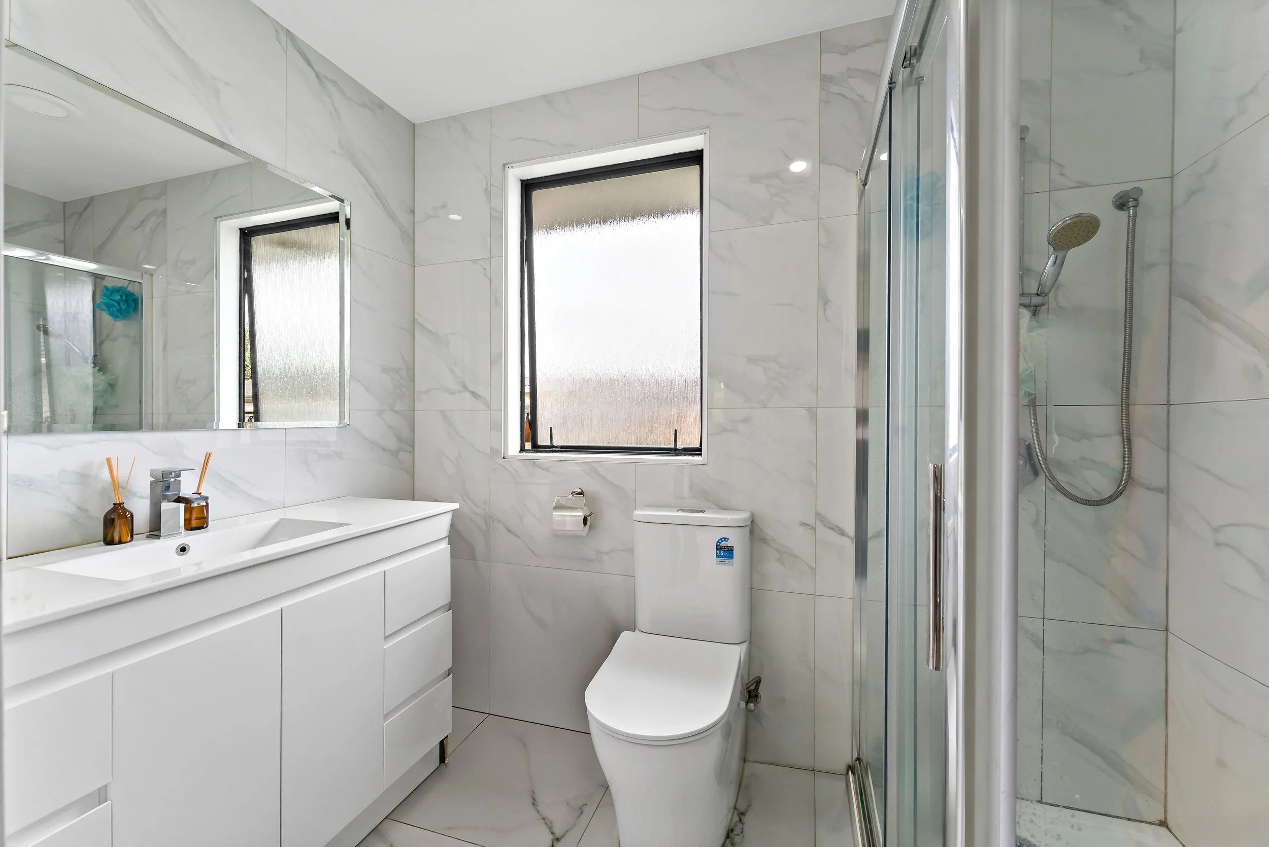 Modern bathroom with white marble walls and floor, a white vanity with a mirror, a frosted window, toilet, and glass-enclosed shower.