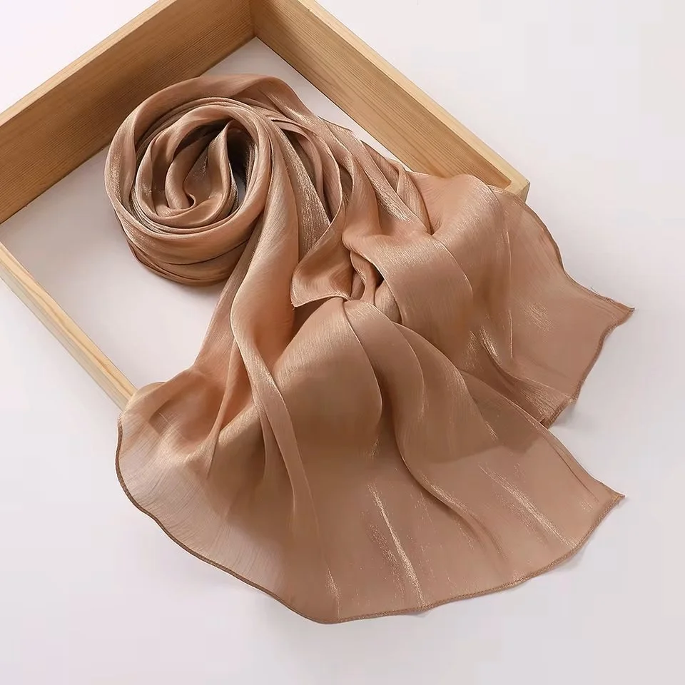 A sheer, satin scarf in a taupe color, draped and partially rolled, placed inside a light wooden box against a white background.